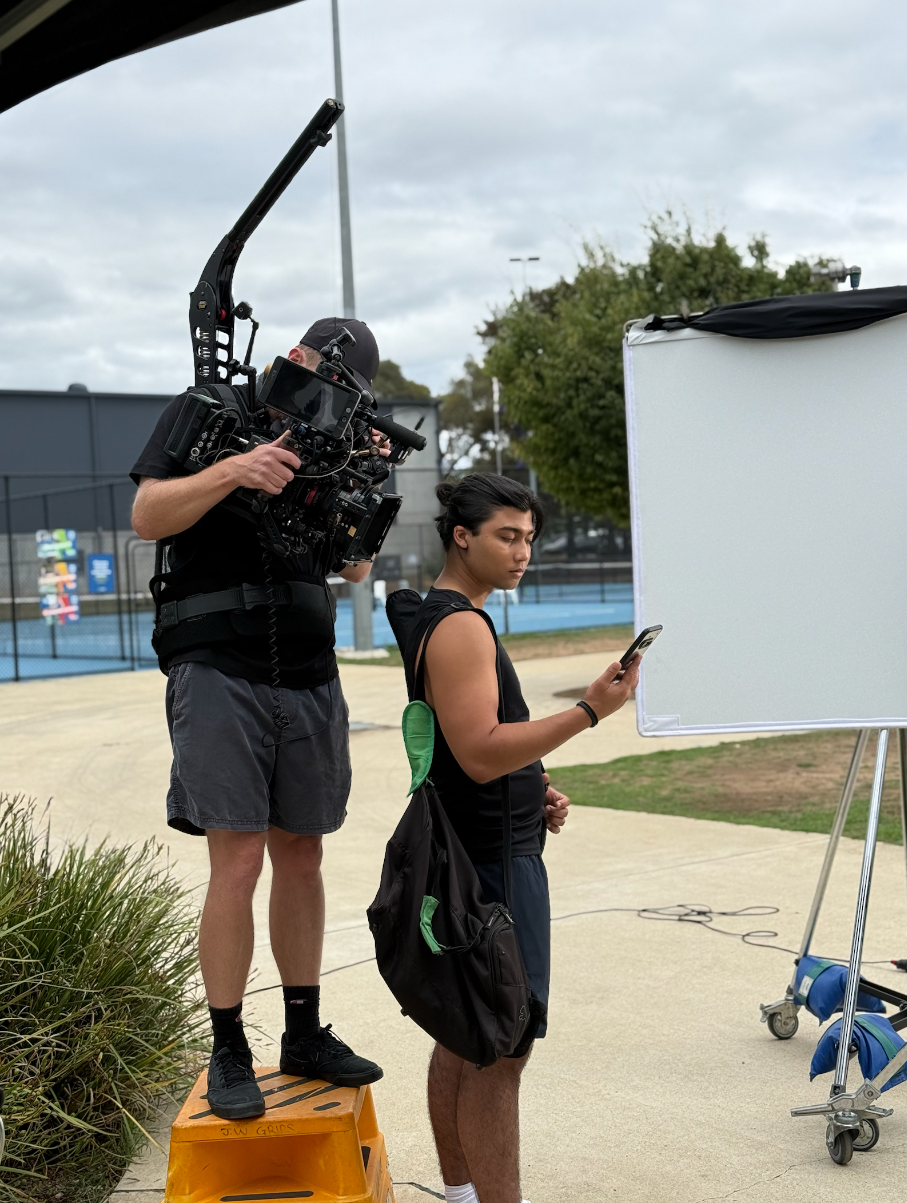Camera operator adjusting equipment on a person using a smartphone outdoors near a tennis court, with a large reflector or diffuser to the right.