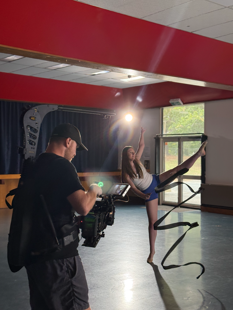 A photographer films a dancer practicing with a harness in a studio with large windows and red ceiling beams.