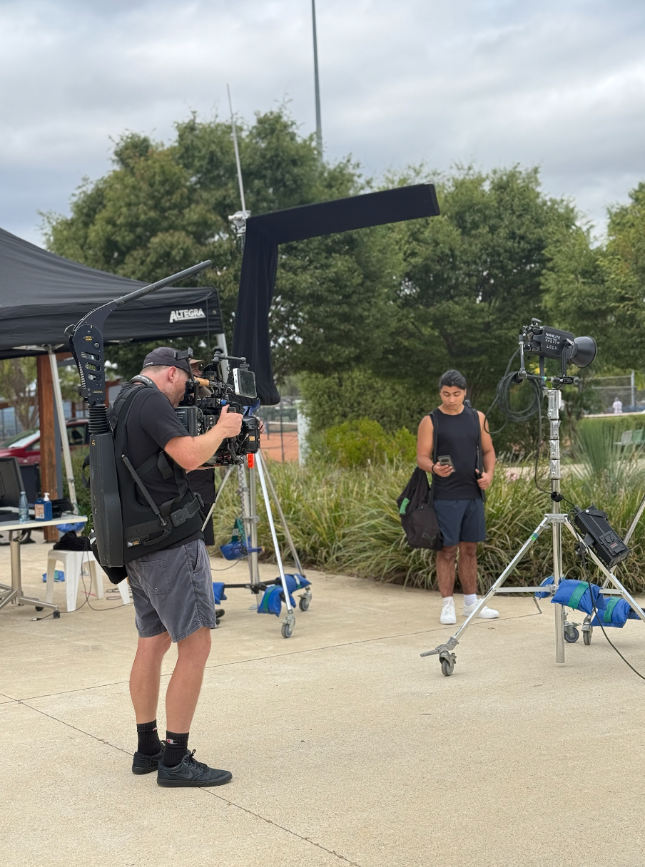 A person operating a video camera on a tripod captures a young man standing in front of a camera, outdoors near greenery and a sports field.