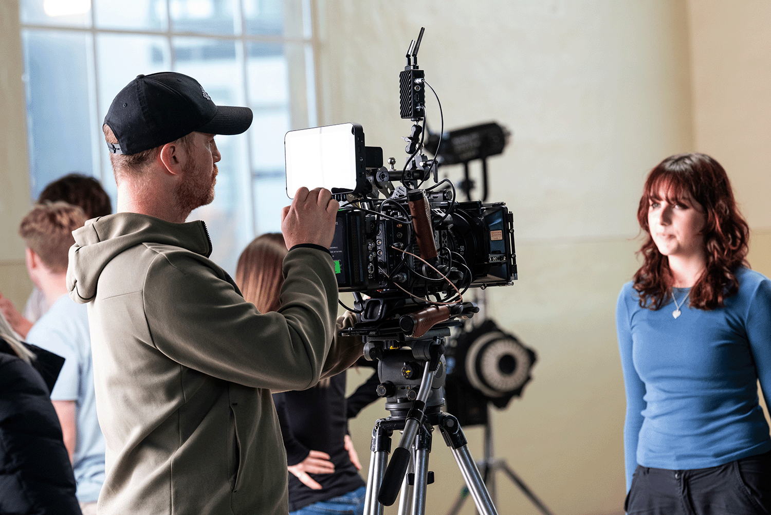 A cameraman filming a woman in a blue shirt during a video recording session or interview in an indoor setting with other people in the background.