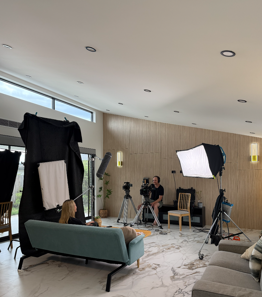 A woman is sitting on a mint green sofa in a modern indoor setting with filming equipment and crew around her, including cameras, lights, and sound equipment.