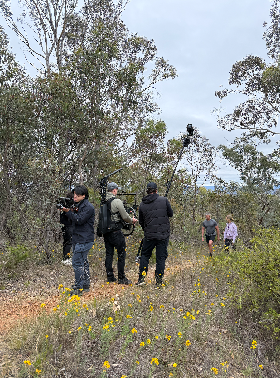 Filming crew with cameras and equipment filming two people walking through a wooded area with yellow flowers.