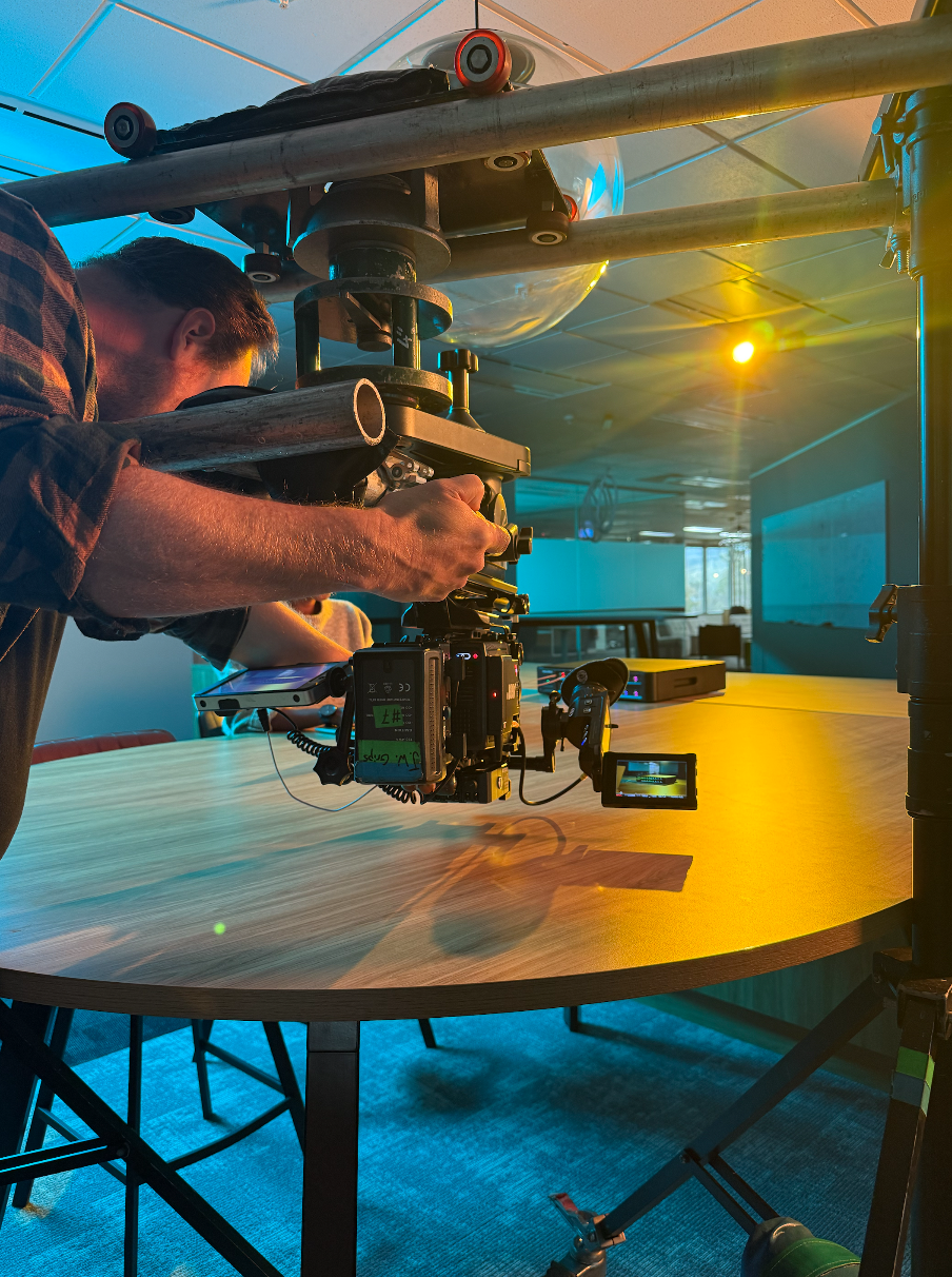 A man operating a camera mounted on a mechanical rig over a round conference table in a modern room with large screens on the walls, with colorful lighting in the background.