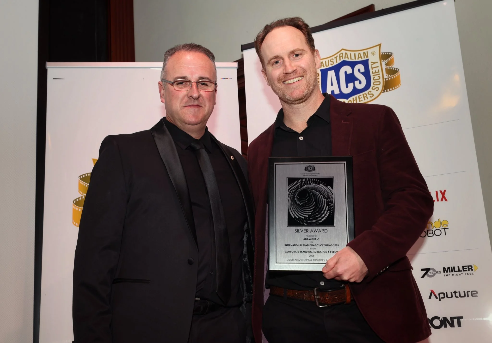 Two men in formal attire standing together at an award ceremony. One man is holding a plaque that reads 'Silver Award' and mentions Adam Grant. The background features logos and banners from Australian awards and sponsors.