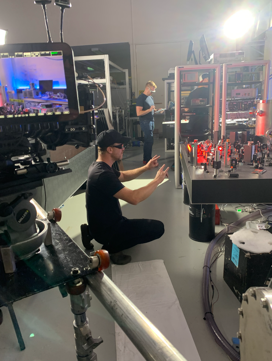 Two technicians working in a laboratory with scientific equipment and laser setup, one kneeling and giving signals, the other standing and reading a clipboard.