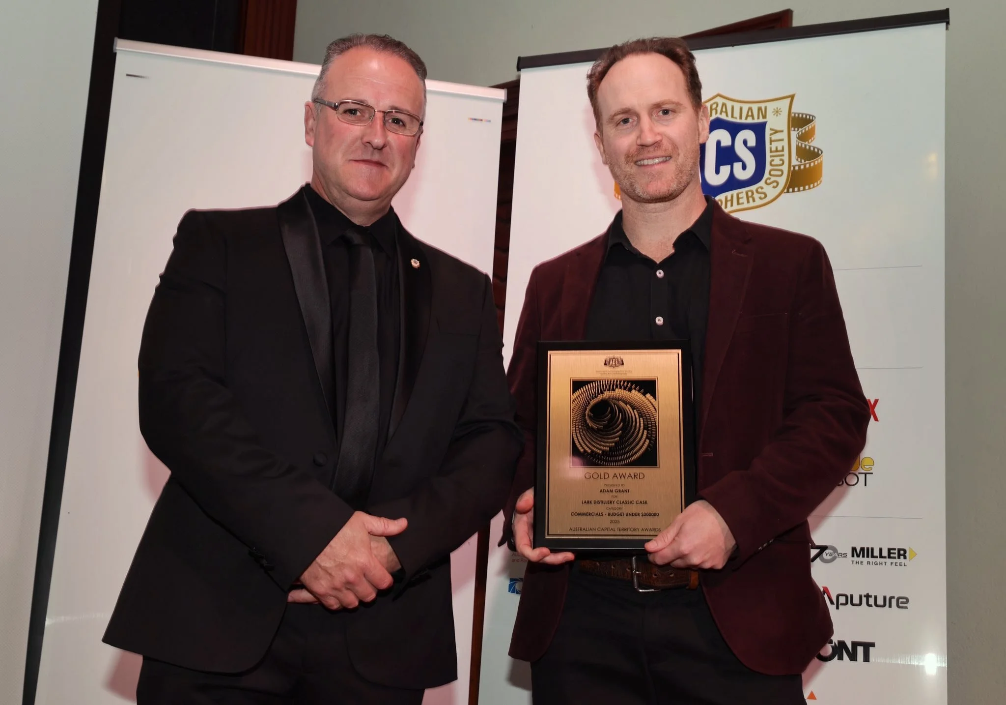 Two men in suits holding a plaque at an award ceremony. Behind them are banners with logos and text.