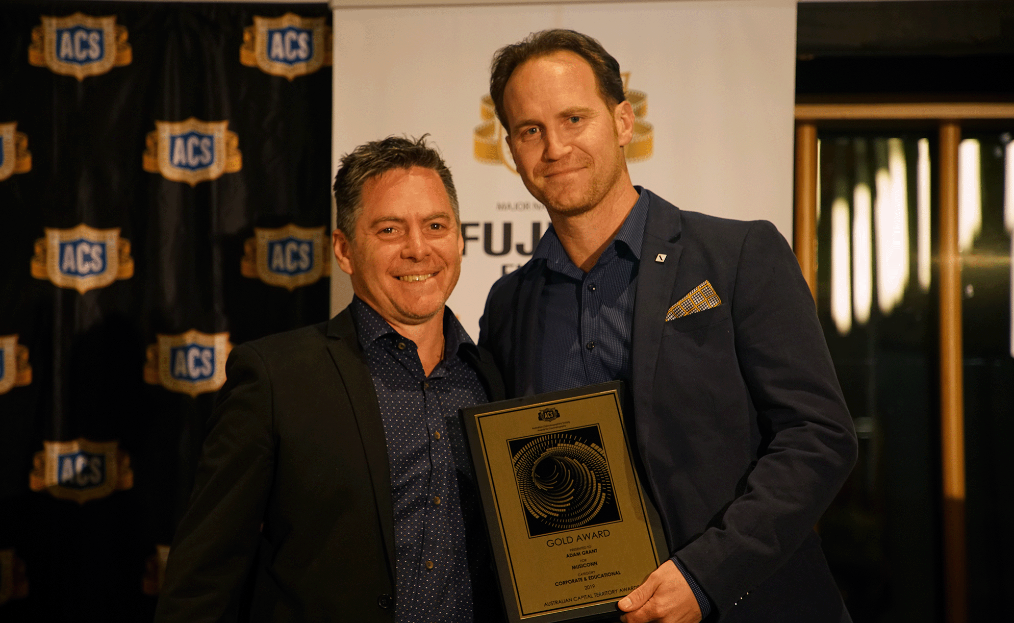 Two men in formal attire standing together at an award ceremony, one holding a gold award plaque. The background features a black curtain with multiple ACS logos and a white banner with a gold emblem.