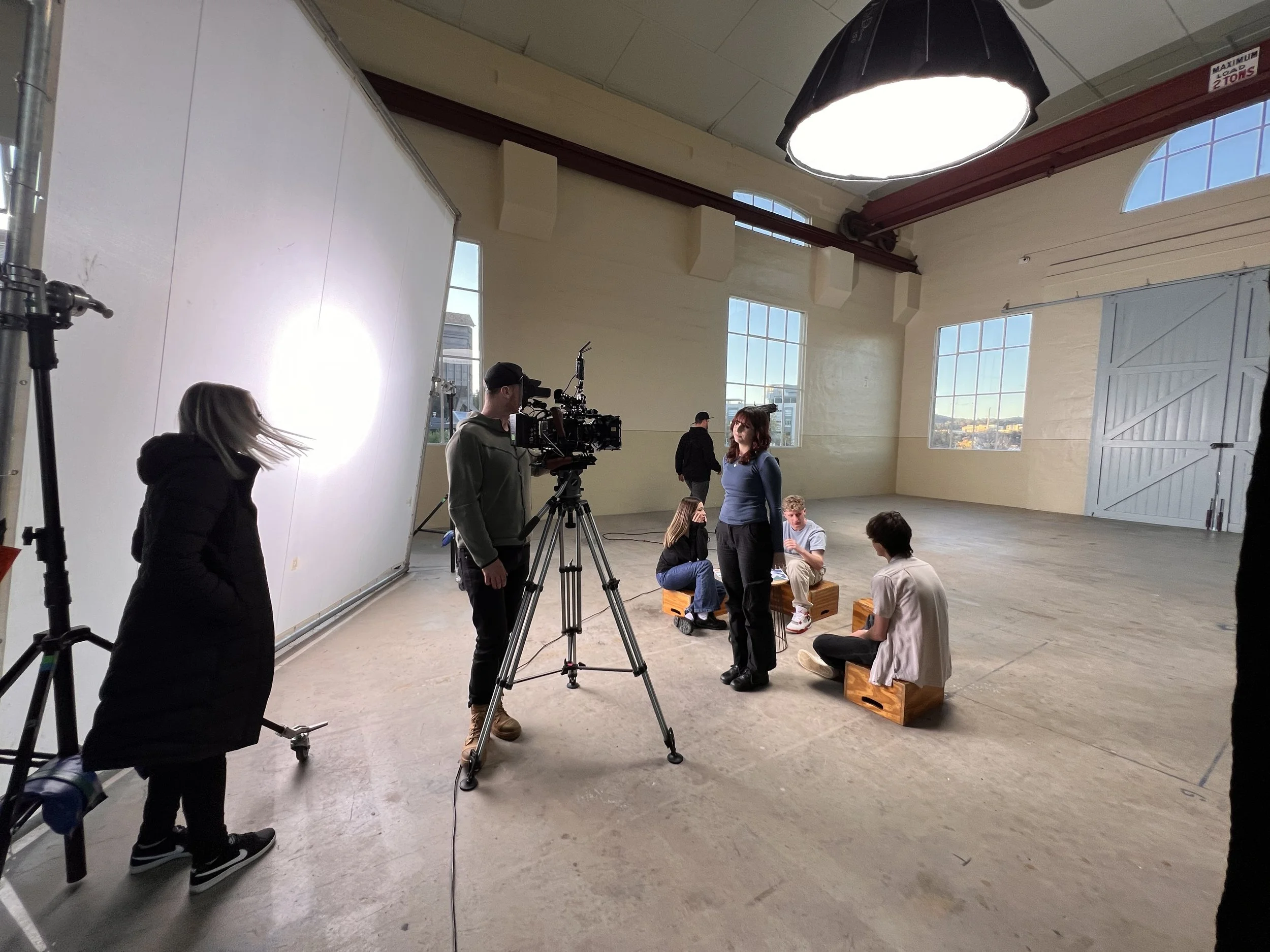 Film crew setting up a scene in a spacious studio with large windows, showing actors sitting and standing during a rehearsal or filming process.