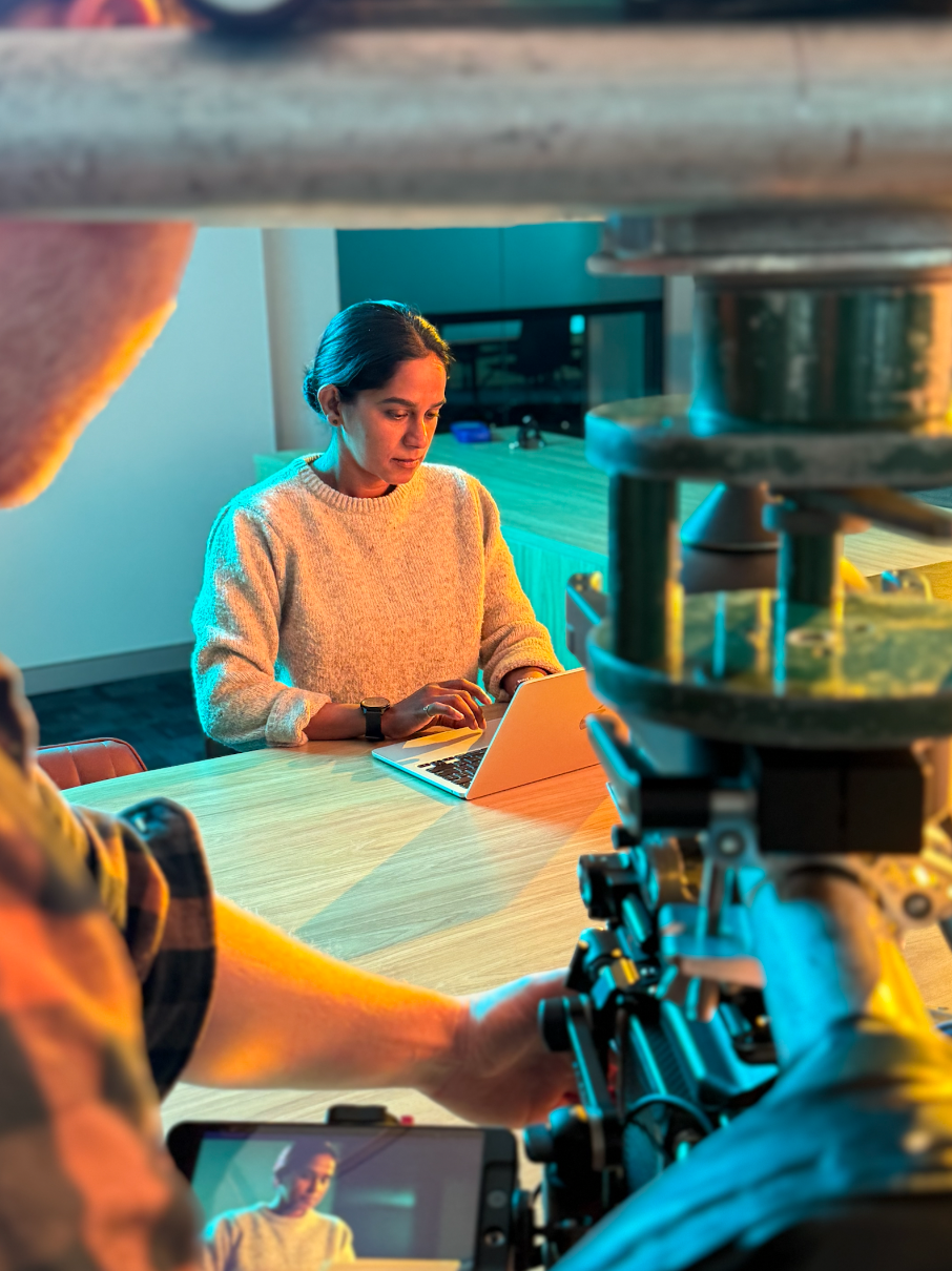 A woman in a light-colored sweater sitting at a table and working on a laptop, with a filming crew recording her using a professional camera and a smartphone.