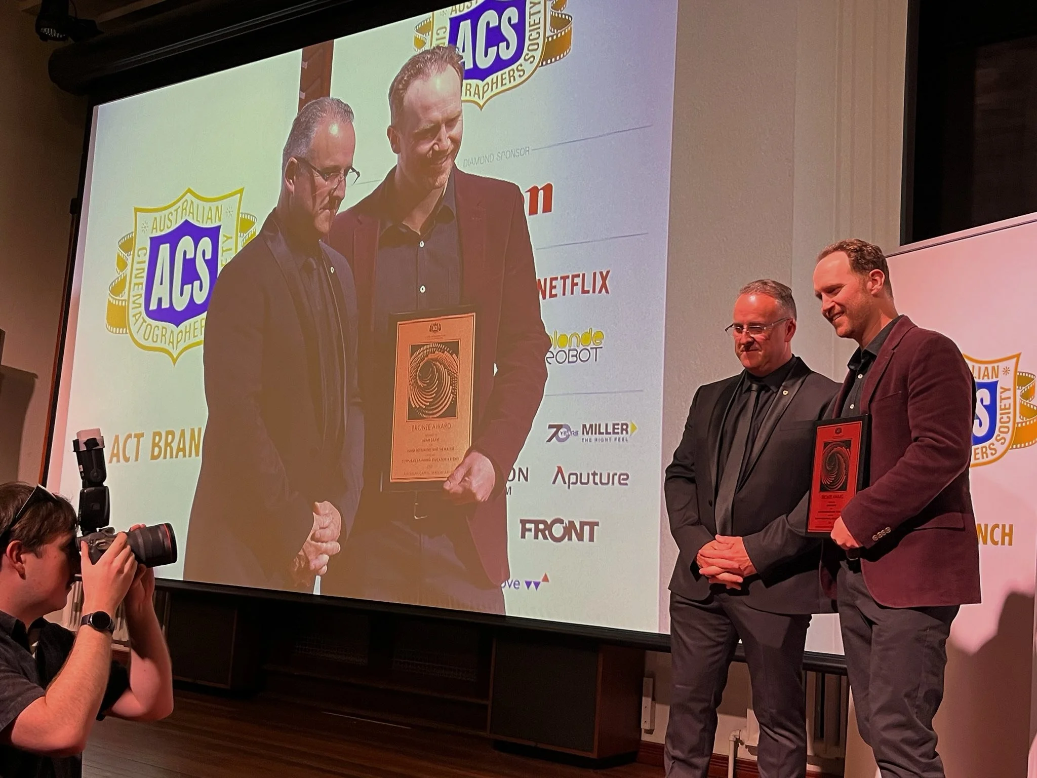 Three men in suits standing on a stage, receiving an award, with a large projector screen behind them displaying event logos and sponsors.
