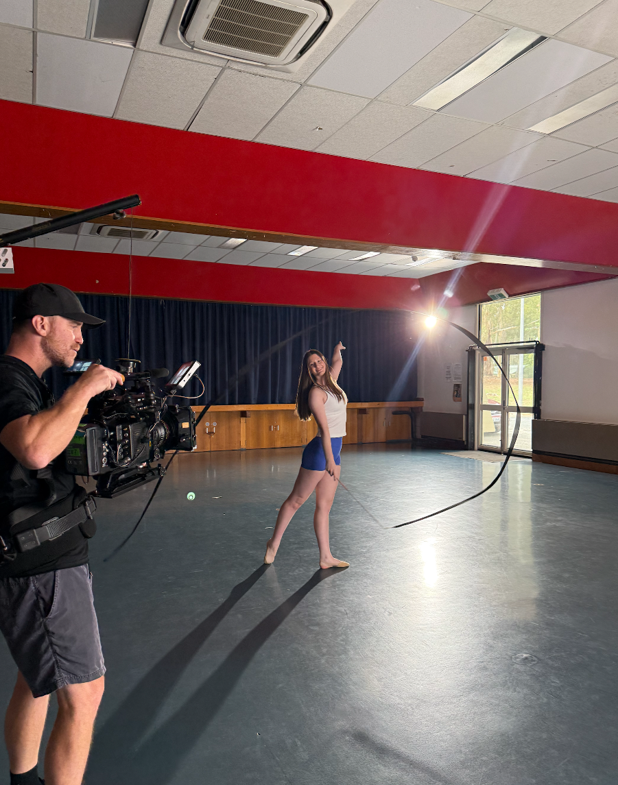 A young woman in a white tank top and blue shorts practicing with a bow and arrow indoors, in front of a camera operator filming her. She is smiling and posing, with a dark blue curtain and a red stripe on the wall behind her, and sunlight coming thr