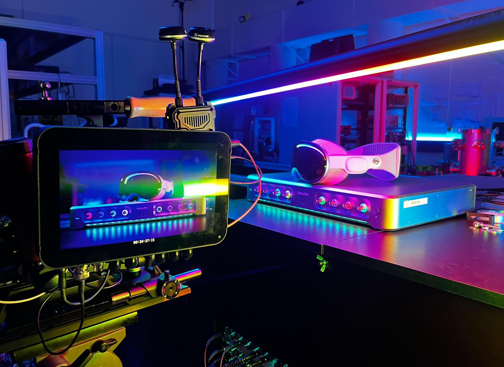 A digital setup with a VR headset on a workbench, a 3D printer, and a tablet showing a similar scene, illuminated by colorful LED lights in a tech workspace.