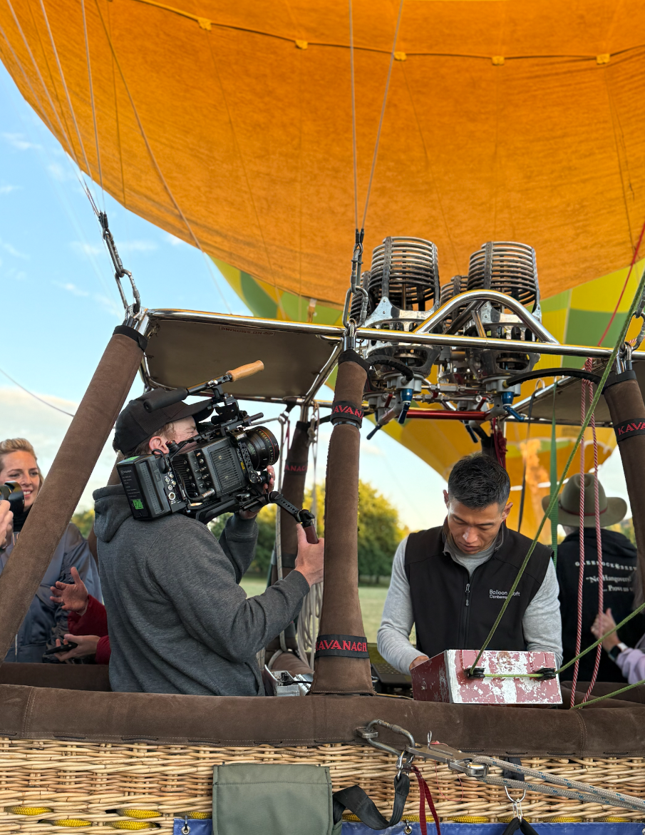 People preparing a hot air balloon for flight; a cameraman filming the scene, with one man adjusting the propane burner.