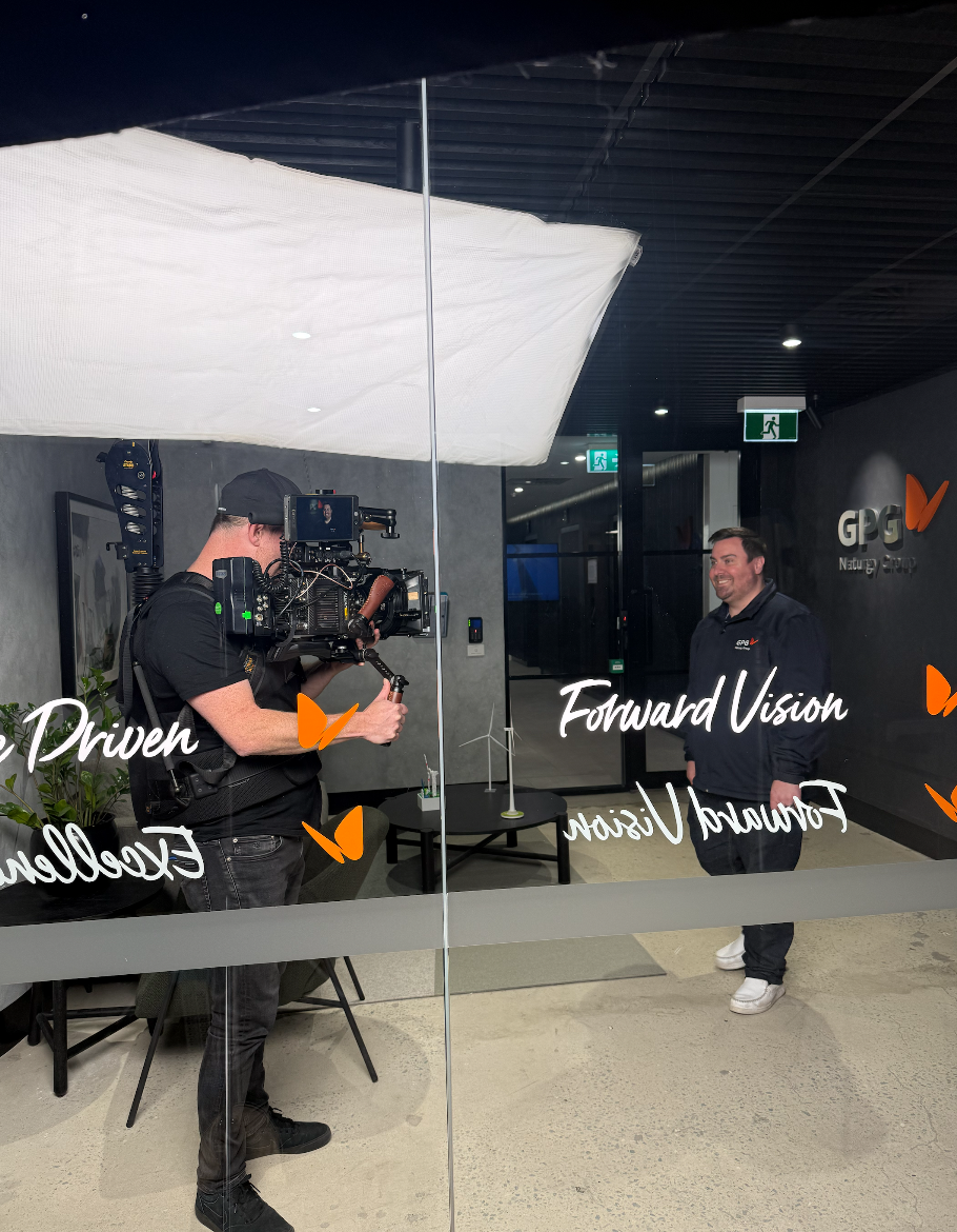 A man being filmed by a camera operator inside an office with glass walls. The man is smiling, wearing a dark hoodie. Text on the glass includes 'Forward Vision' and 'Driven.'