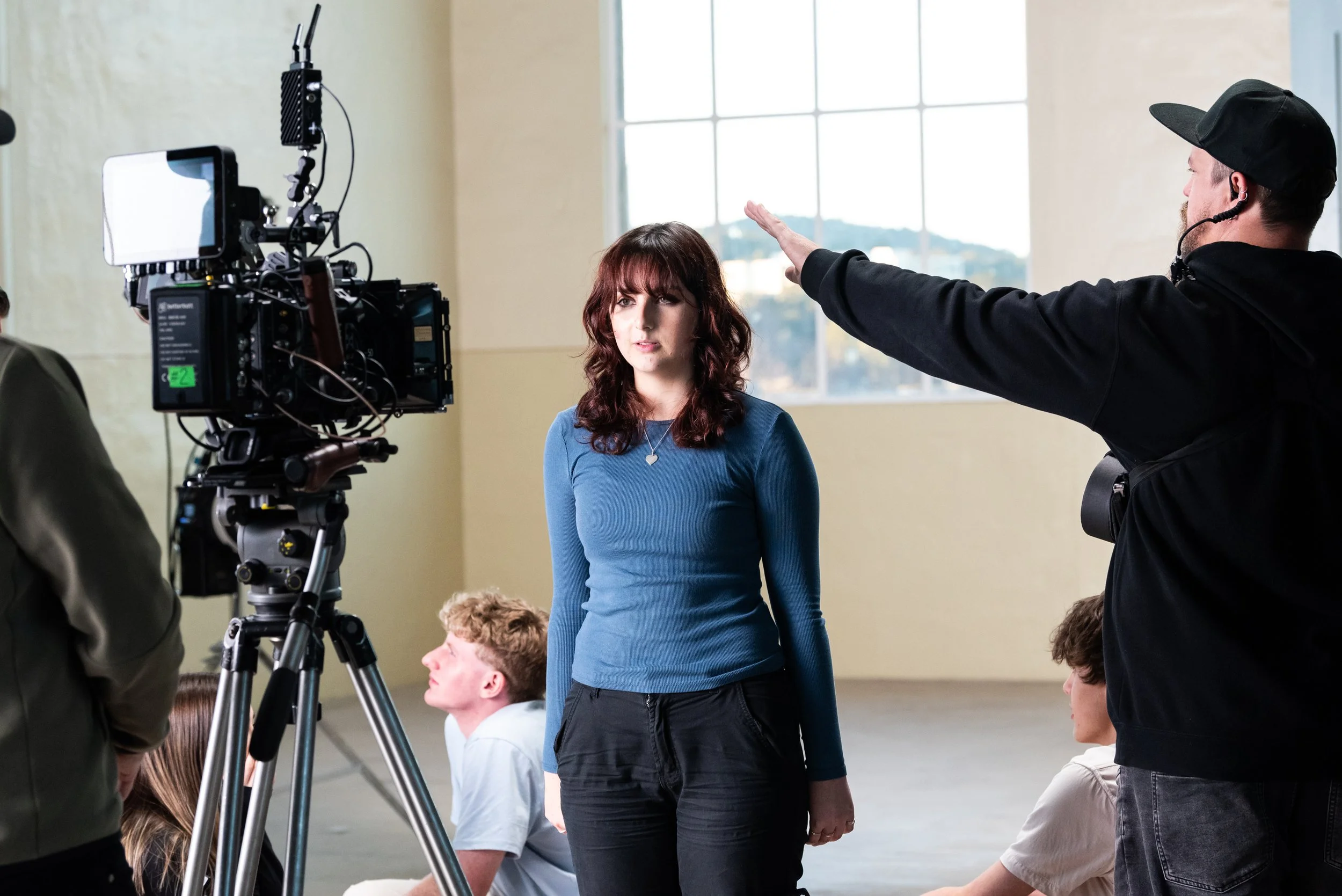 A woman with curly brown hair standing in front of a camera being directed by a man in black with a headset, with people seated on the floor around her in a large, sunlit room.