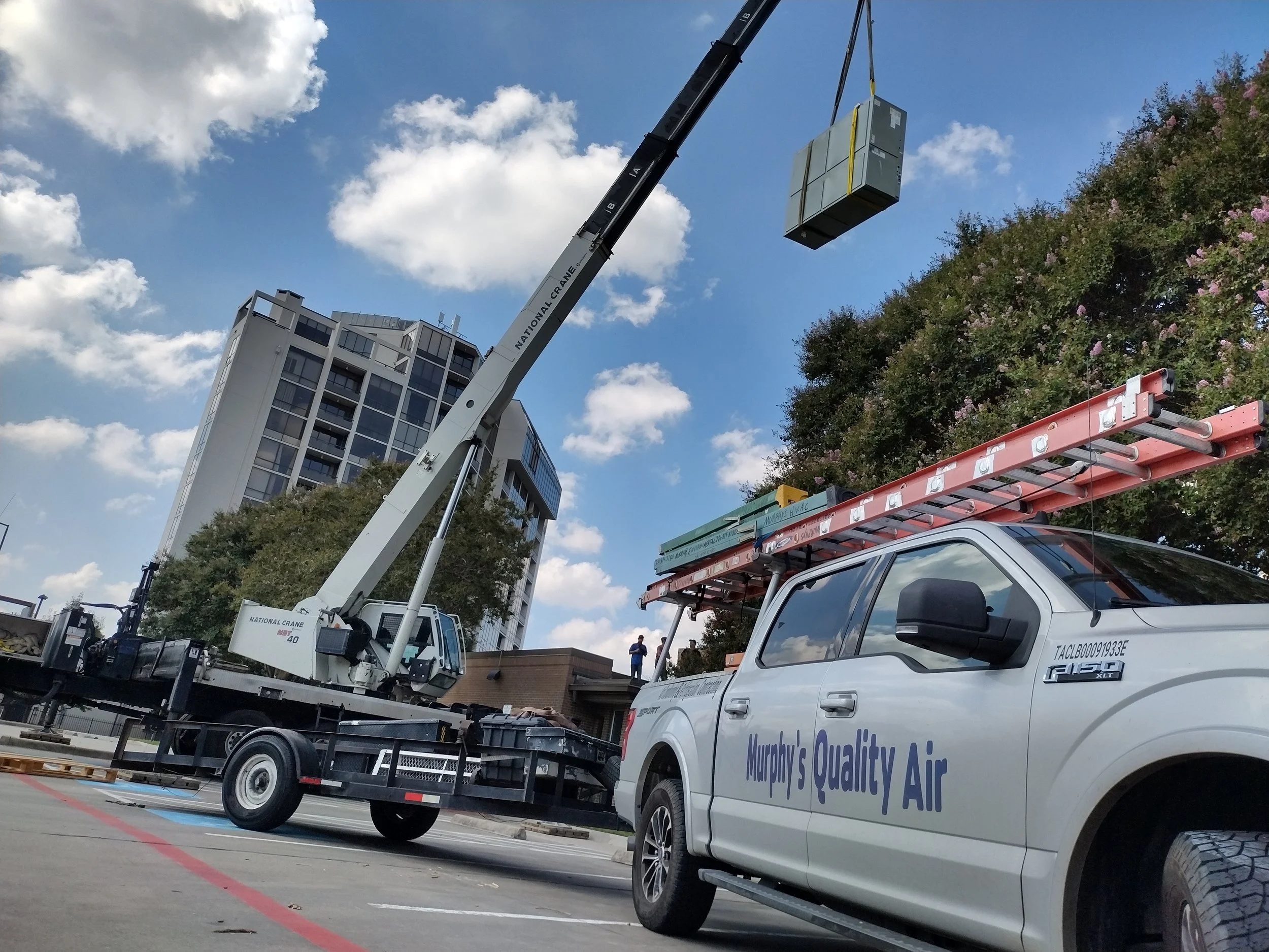 Crane hoisting HVAC unit next to white truck with ladders on top and "Murphy's Quality Air" logo, building and trees in background.