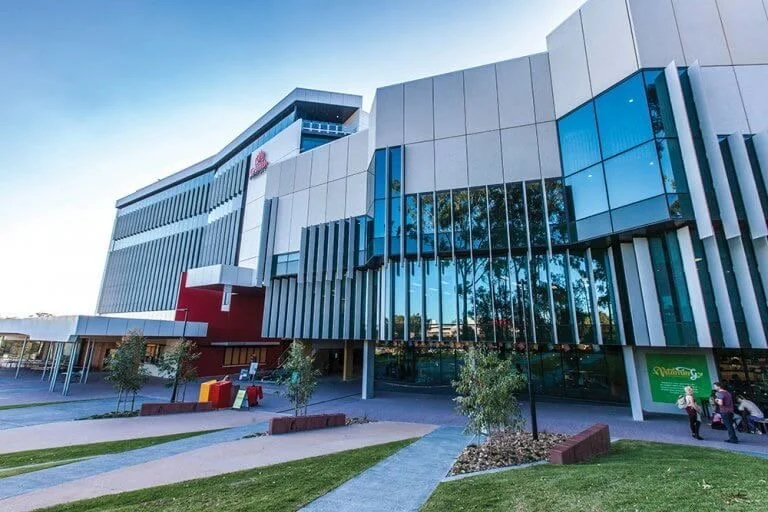 Modern multi-story building with glass windows and vertical metal accents, trees in the landscaped foreground, and a few people walking nearby.