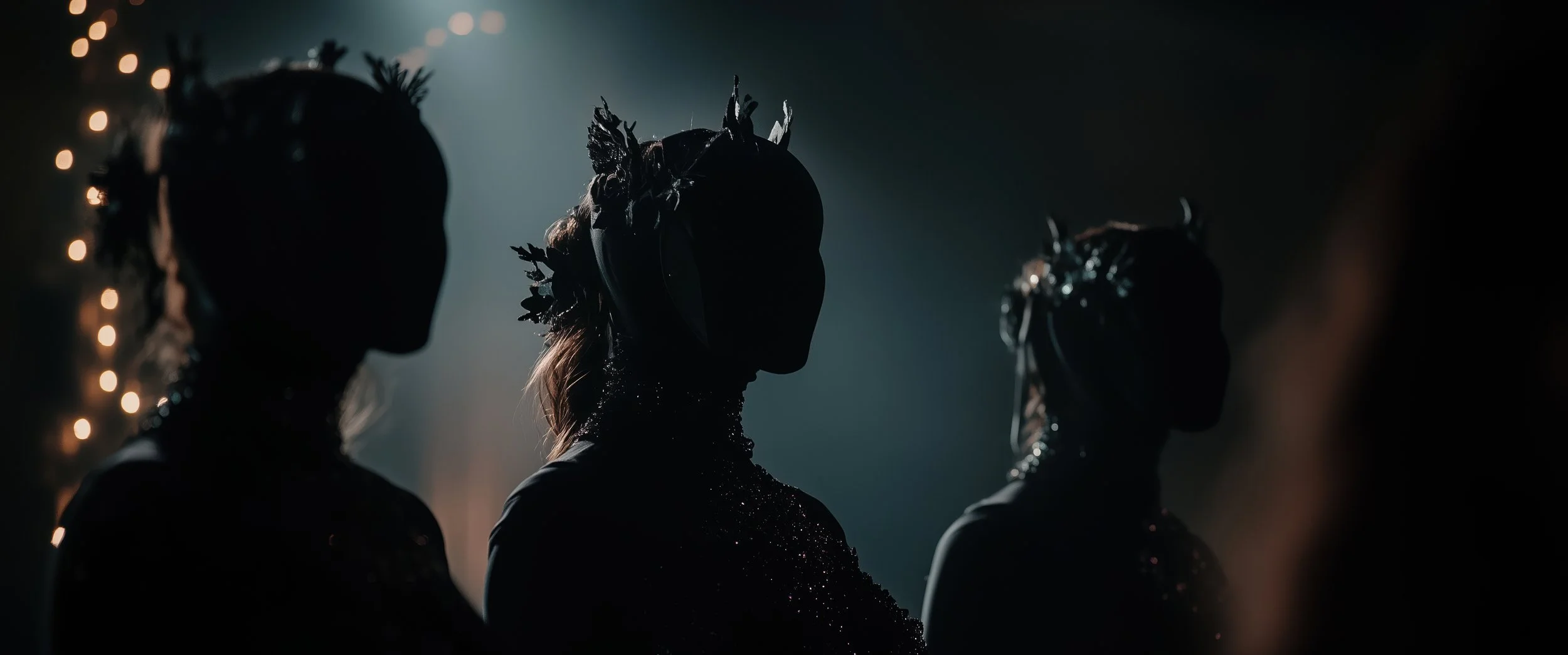 Silhouettes of three women wearing dark, ornate dresses and floral crowns, standing in a dimly lit setting with blurred lights in the background.