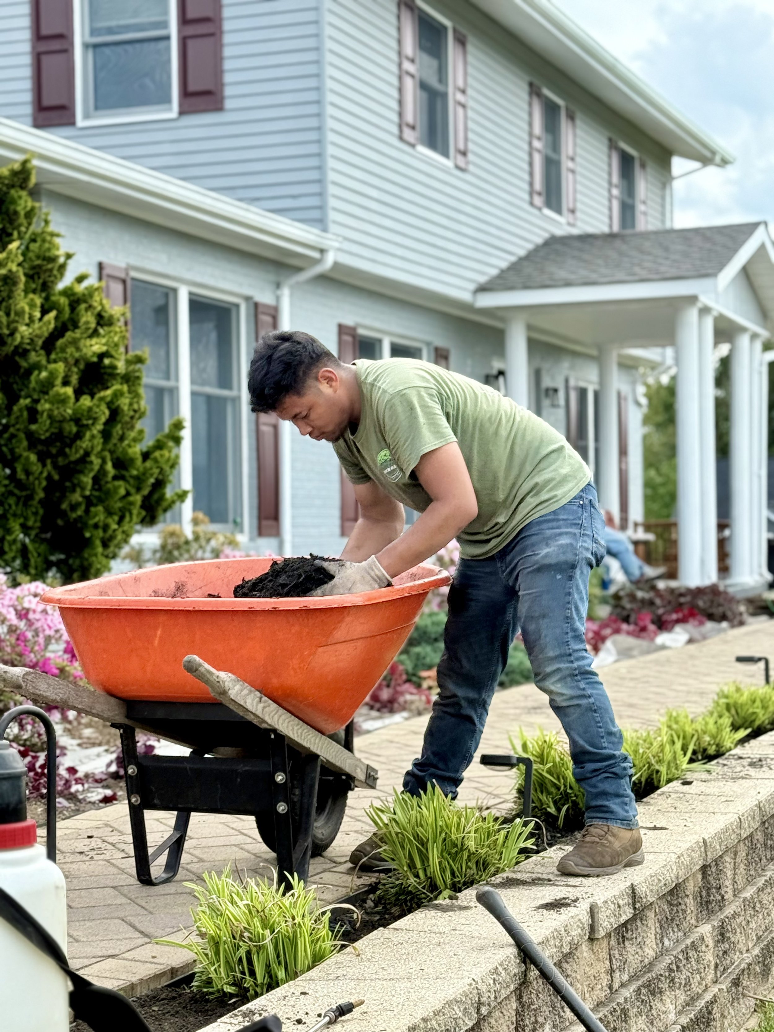 A man installing mulch in a flower bed outside a two-story house with blue siding and brown shutters.