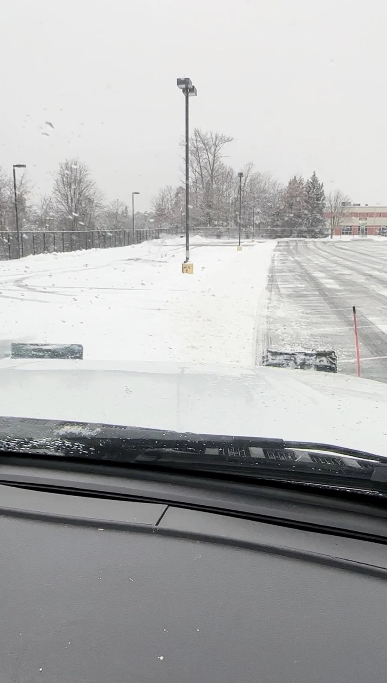 View of a snow-covered parking lot from inside a vehicle, with snowplow equipment clearing the parking space, snow falling, and trees in the distance.