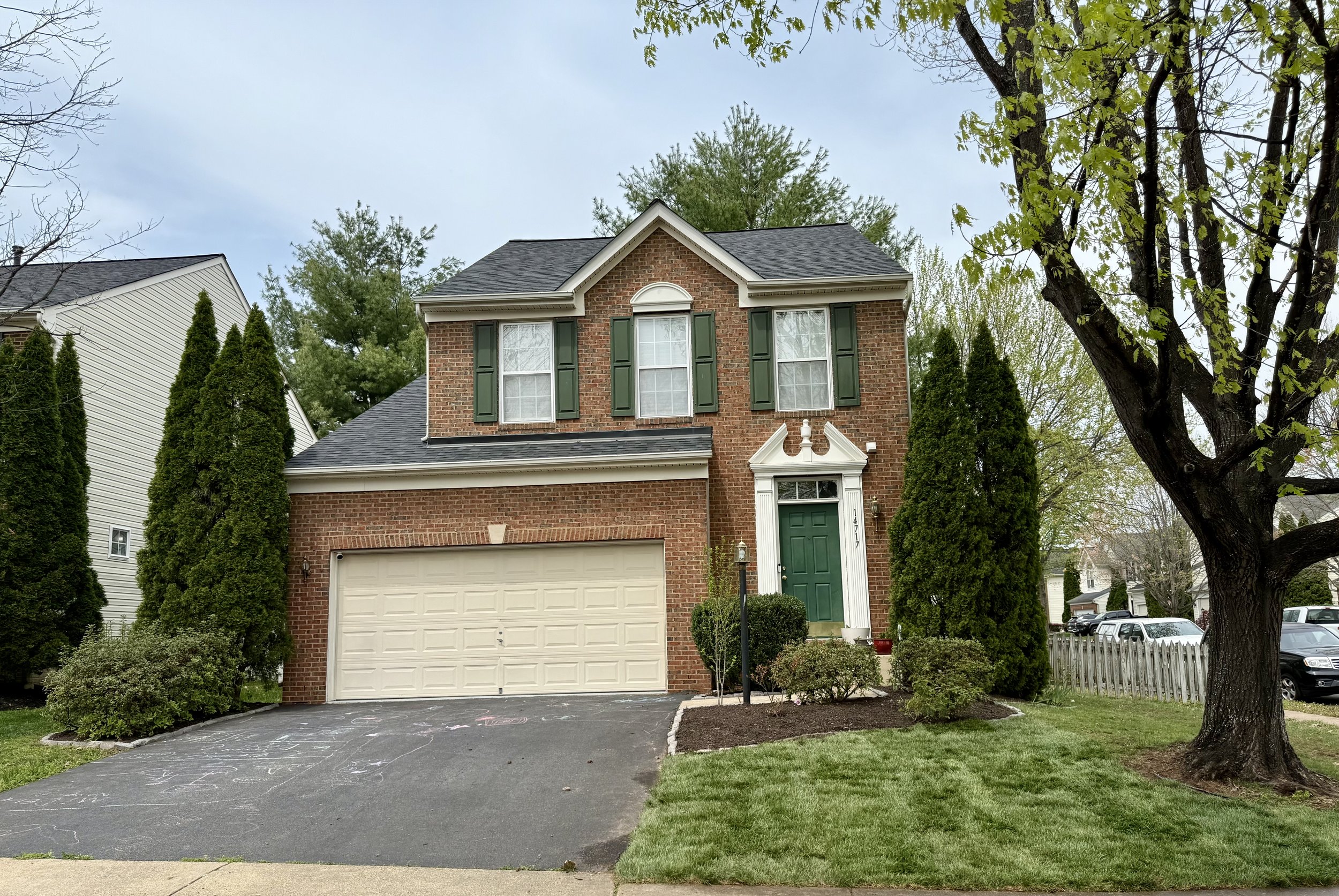 A two-story brick house with green shutters and a green front door gated by white trim, surrounded by tall evergreen trees and a well-maintained lawn, with a driveway in front.