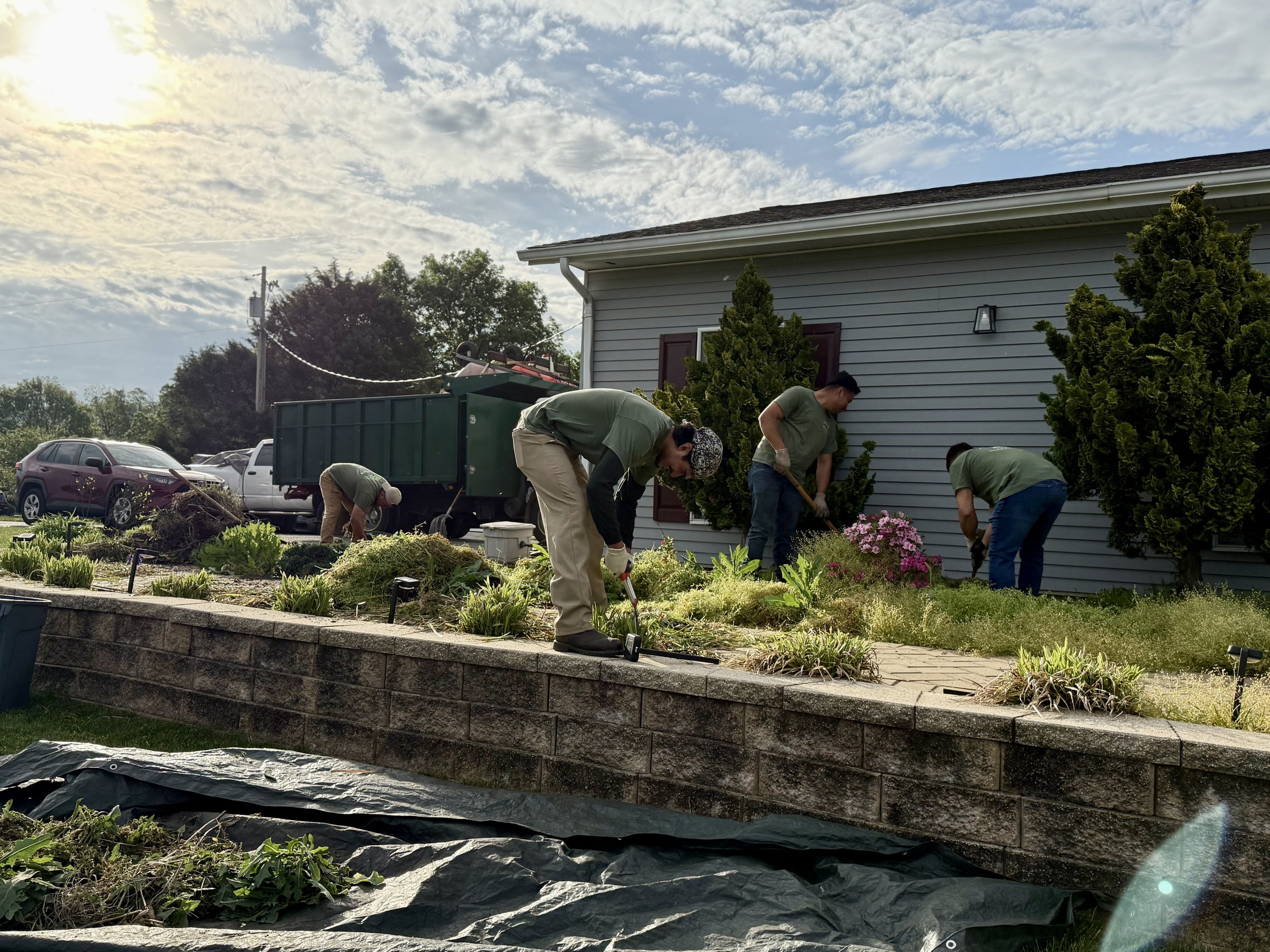 A group of five people are working together to landscape a garden area outside a house, with some using shovels and others using rake or hand tools, under a partly cloudy sky in the late afternoon.