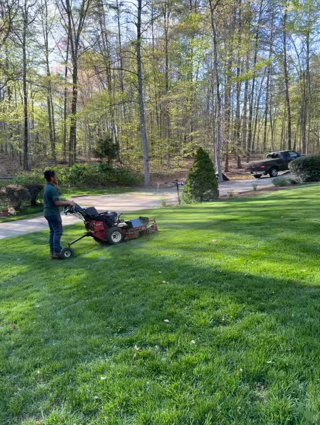 A person mowing a grassy lawn with a riding lawn mower in a backyard, with trees and parked cars visible in the background.
