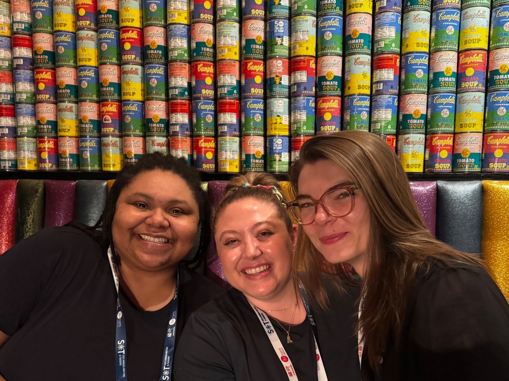 Doctors Forté, Katelyn Polemi, and Rachel Morgan posing together for a photo in Orlando, Florida, at the Society of Toxicology Meeting. Colorful cans make up the background (nod to Andy Warhol).