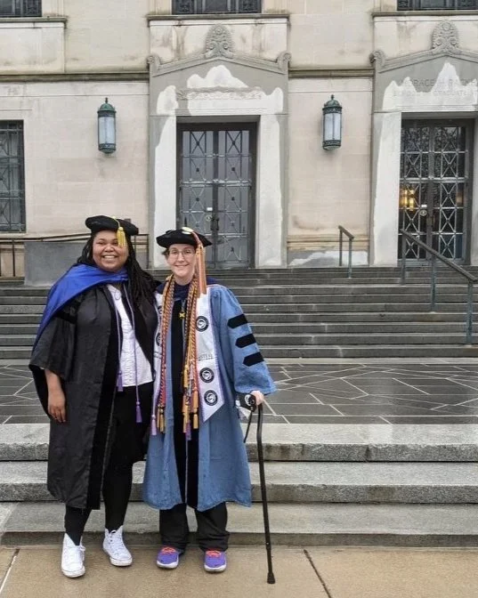 Dr. Forté and Dr. Millar in academic regalia standing in front of a building with steps. One wears a black gown and cap, the other wears a light blue gown and black cap, holding a walking cane. Both are smiling while wearing cords and stoles.