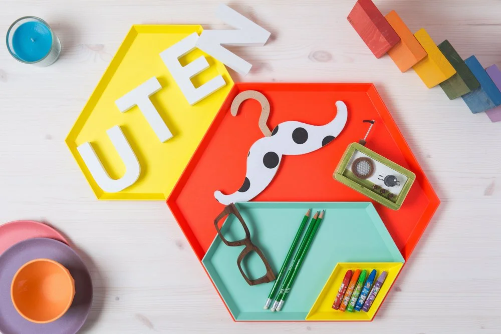 Colorful hexagonal trays with classroom items such as a dollop of blue paint, a paper cutout of a spotted dog, glasses, green pencils, colorful chalk, and a tape recorder, arranged on a light wooden surface.