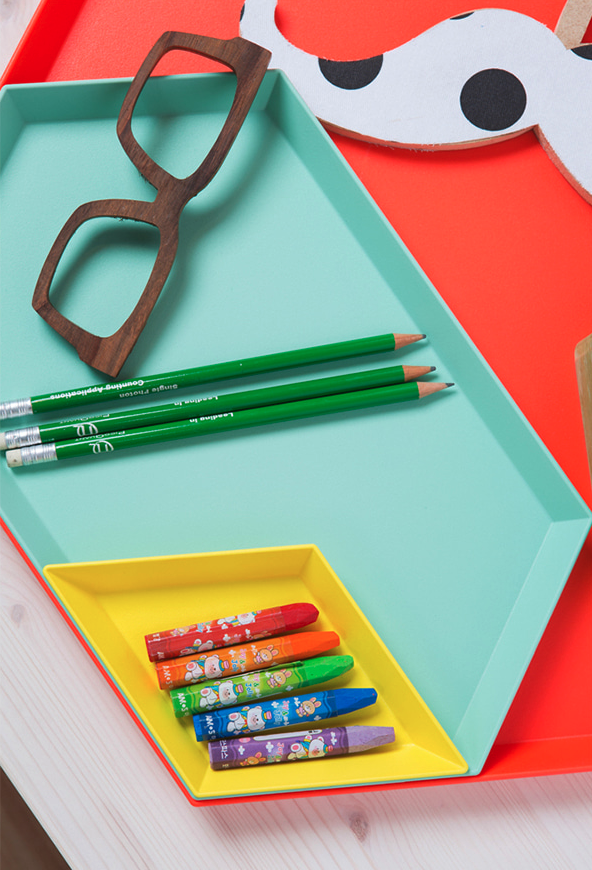 A colorful desk organizer with three green pencils, a pair of black-rimmed glasses, and six Crayola crayons in a yellow compartment, with a red and white paper fox craft on a white wooden surface.