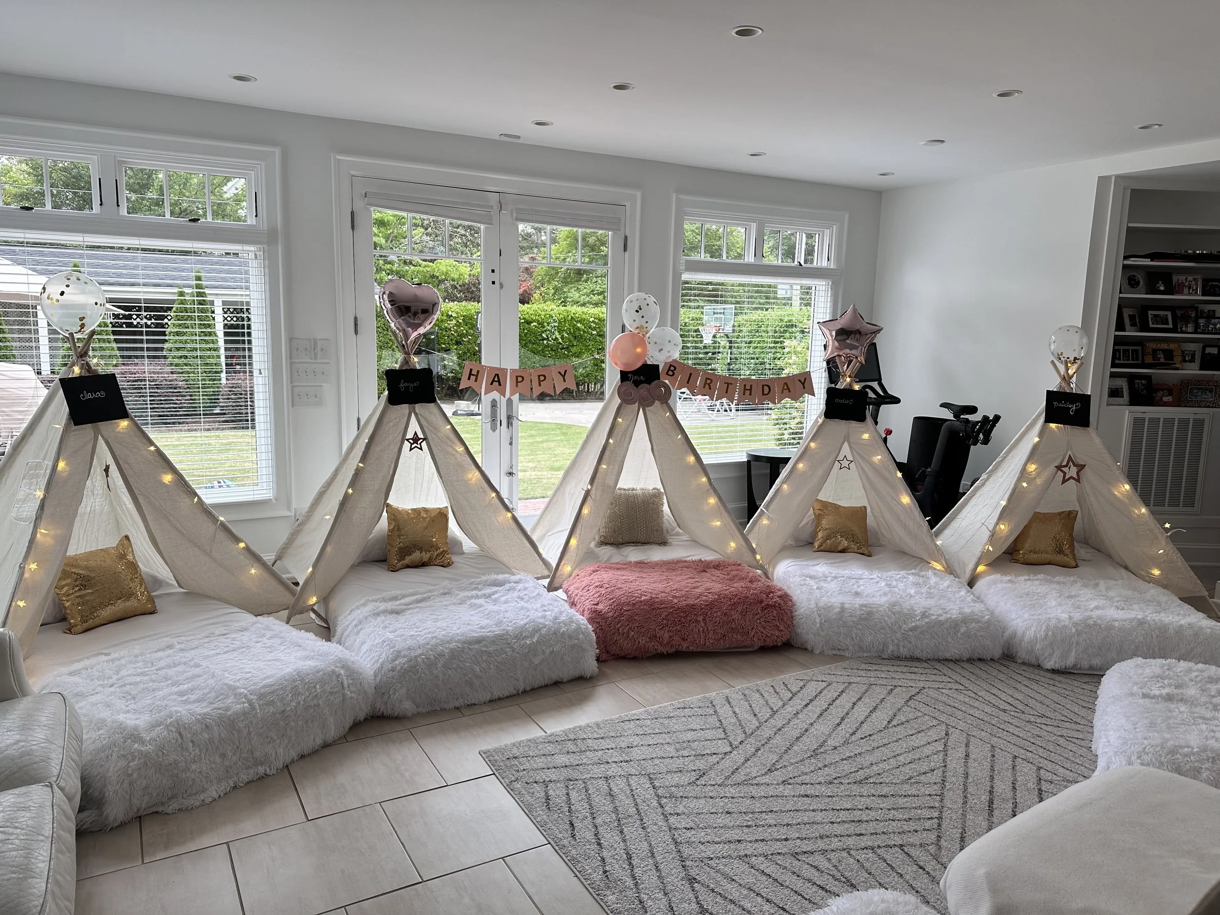 A cozy indoor setup with five small teepees draped with fabric, each adorned with string lights. Inside each teepee are fluffy blankets and decorative pillows. Above, a banner reads 'Happy Birthday.' There are balloons on top of each teepee, and the scene is set in a bright room with large windows.
