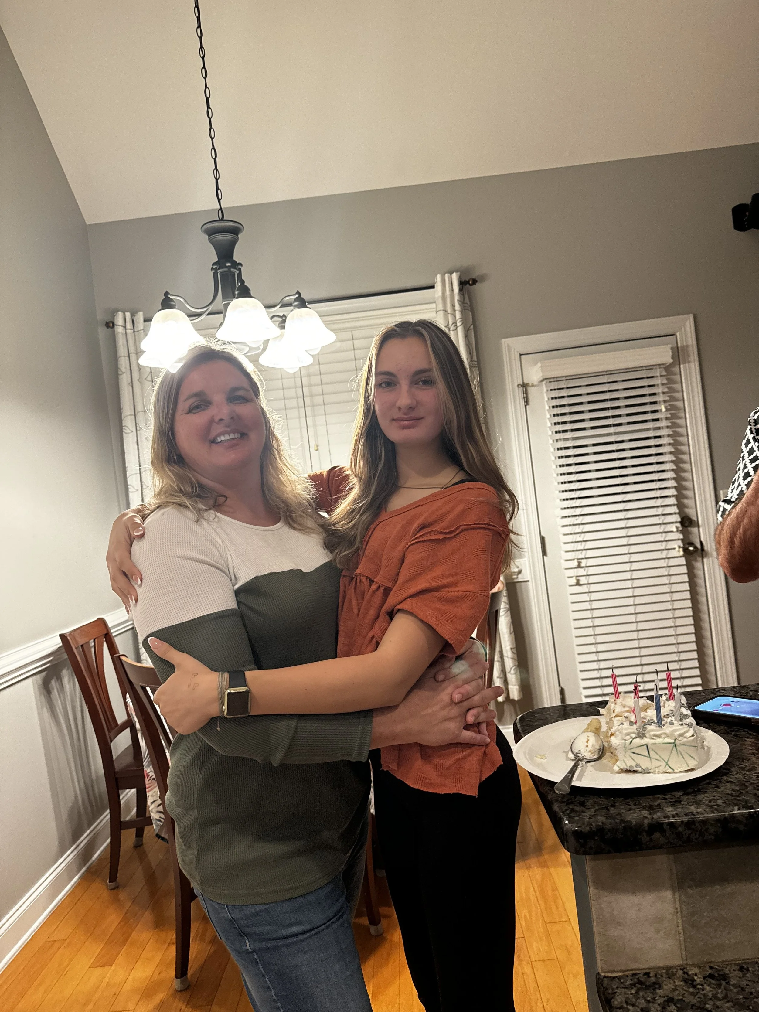 Two people hugging in a kitchen with cake on the counter nearby, decorated with lit candles and a spoon on the plate.