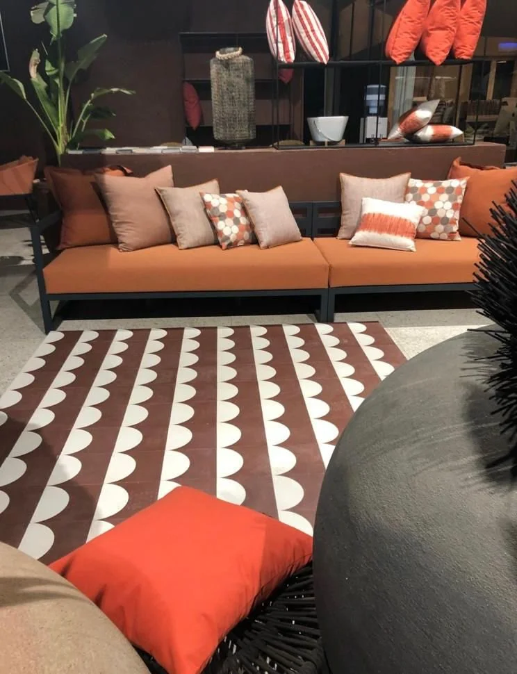 A modern indoor seating area with a long orange cushioned sofa, various throw pillows in beige, orange, and patterned designs, a patterned rug with white semi-circles on a brown background, and decorative shelves with accessories in the background.