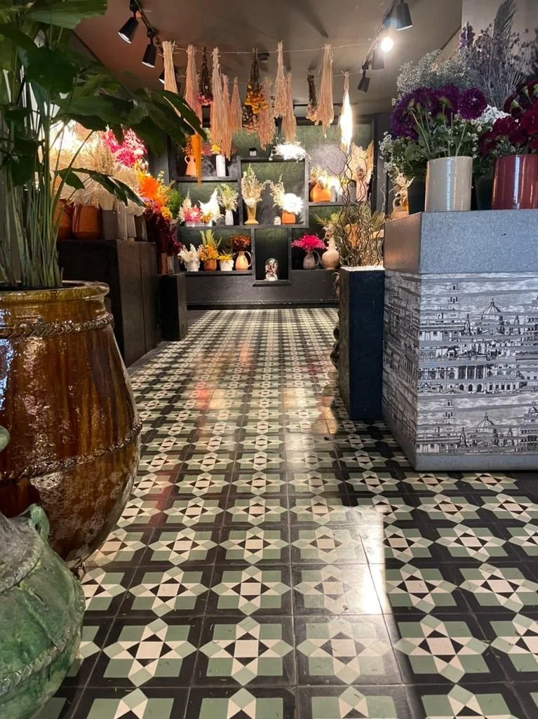 Interior of a flower shop with a patterned tile floor, various potted flowers and plants, and artful displays of flowers on shelves and in vases.