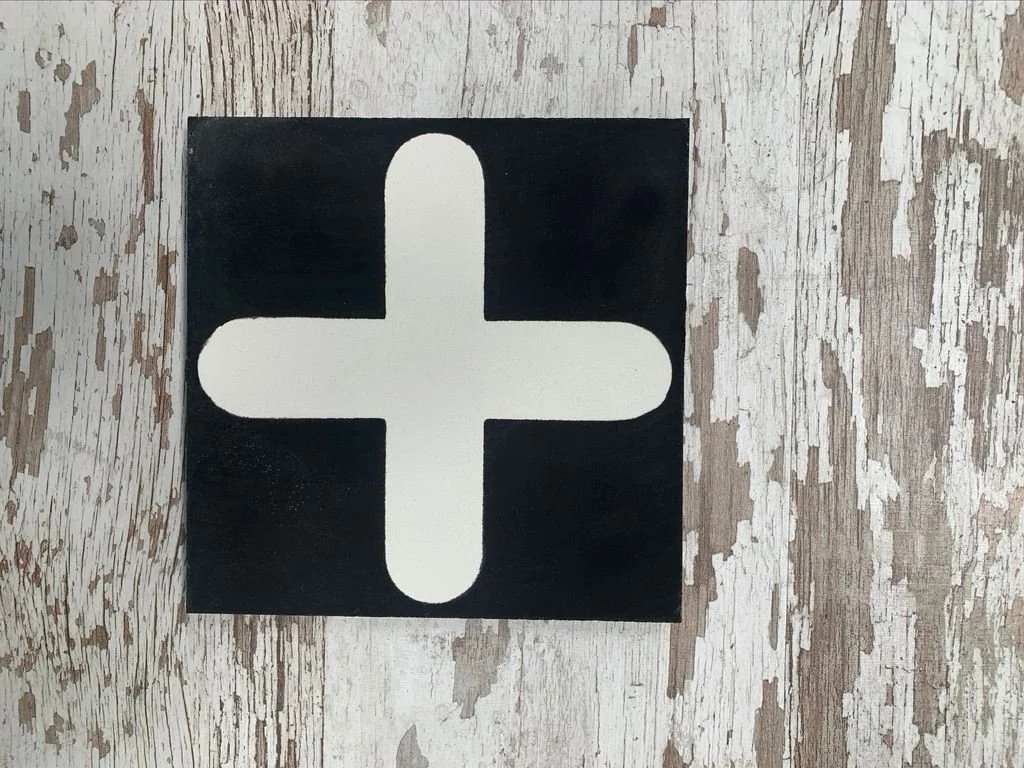 A black tile with a white plus sign in the center, placed on a weathered, white-painted wooden surface.