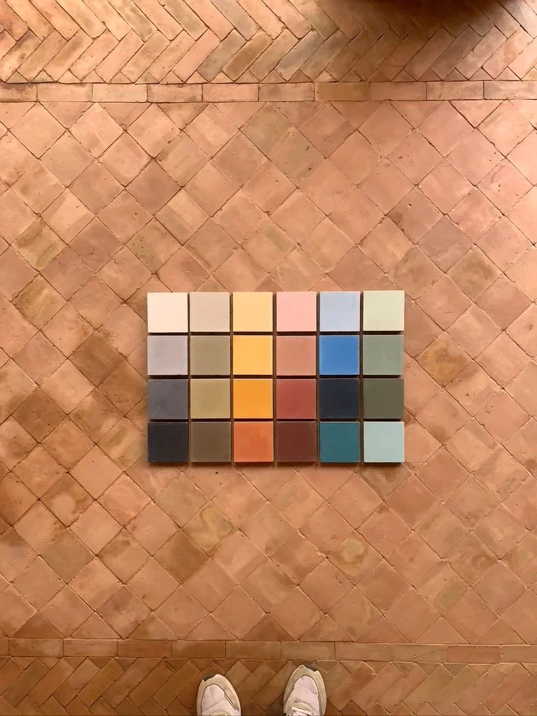 A collection of small square paint color samples arranged in a grid on a wooden floor, with a pair of shoes visible at the bottom of the image.