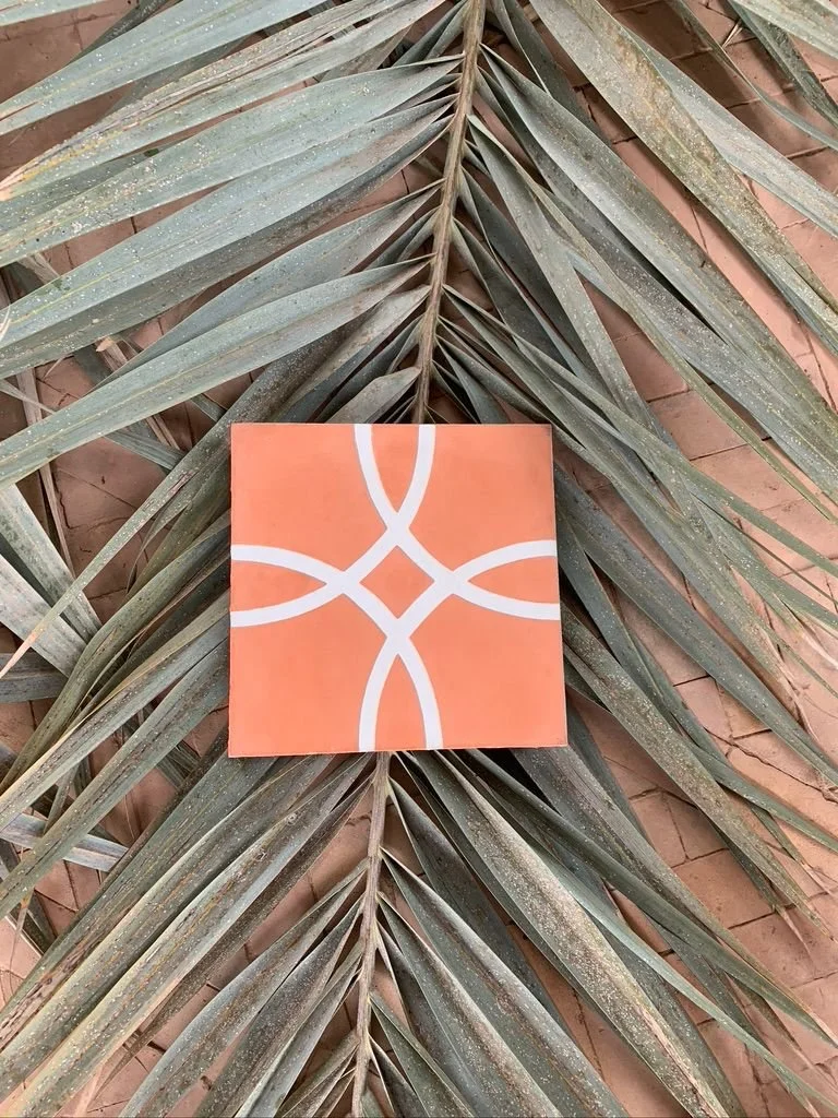 A decorative tile with an orange background and a white, symmetrical curved pattern, placed against a large palm leaf and a brick wall.