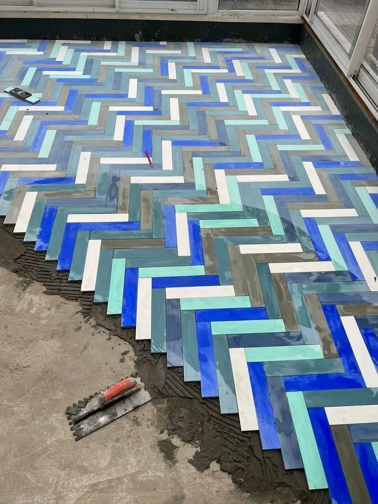 Colorful herringbone pattern of glass tiles in shades of blue, white, gray, and aqua, being installed on a floor near a glass door.