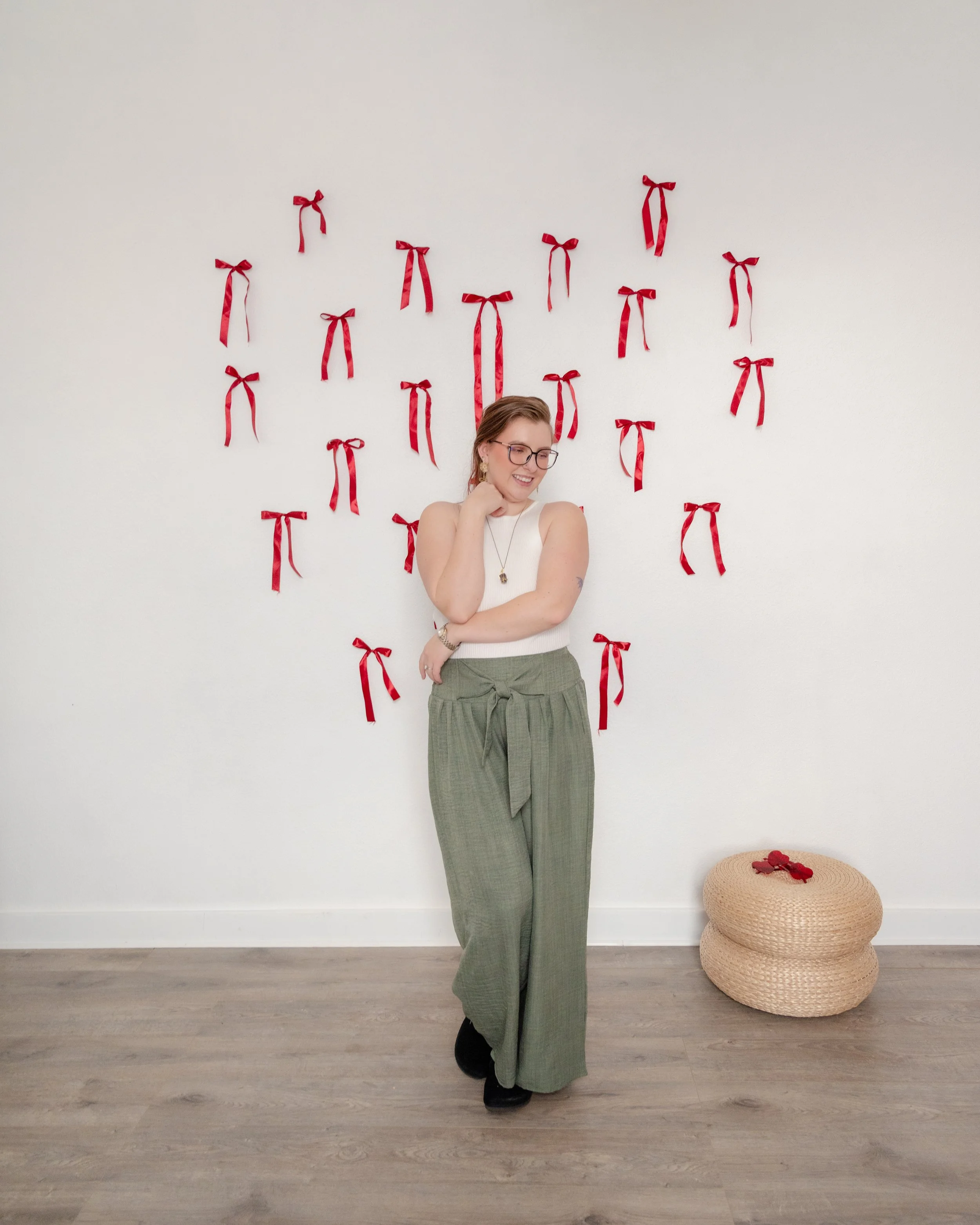 Woman posing in front of a Valentine ribbon wall in The Alcove natural light studio in Springville, Utah, wearing a white sleeveless top and olive green pants.