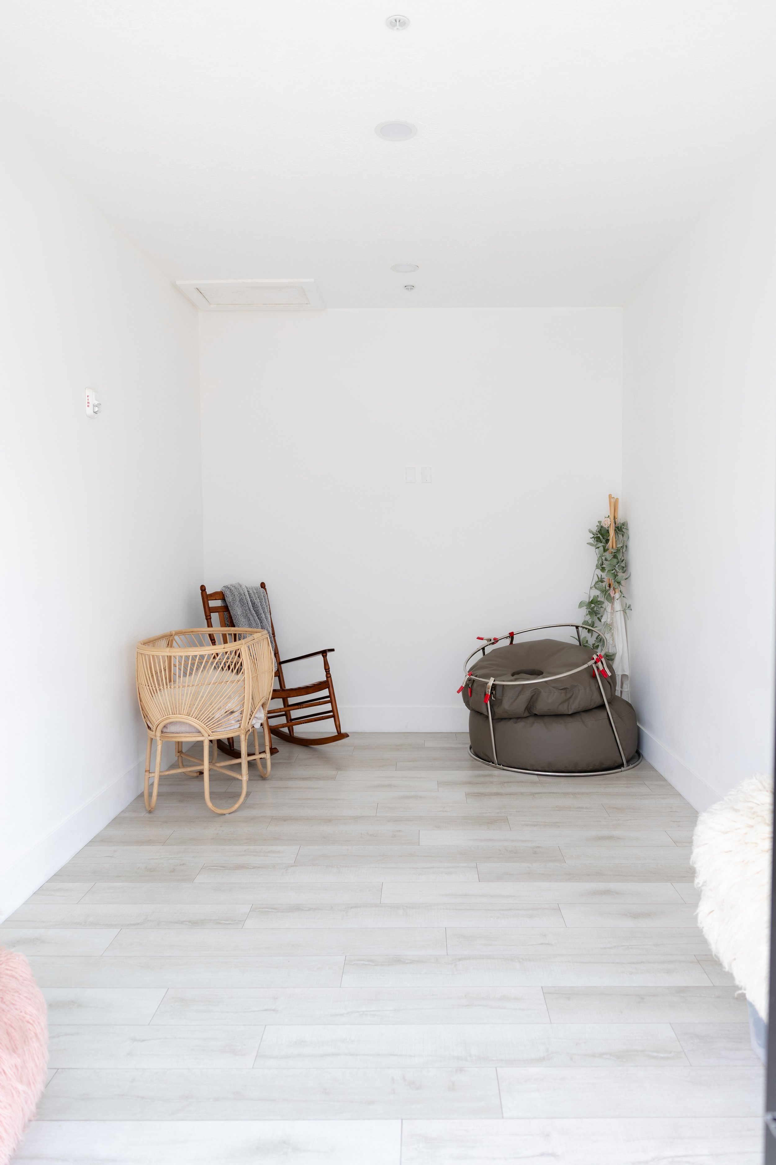 A corner of a room with light wood flooring, a wooden rocking chair, a wicker basket, a round table with a white cloth, and folded colorful blankets and towels stacked on a table and baskets.