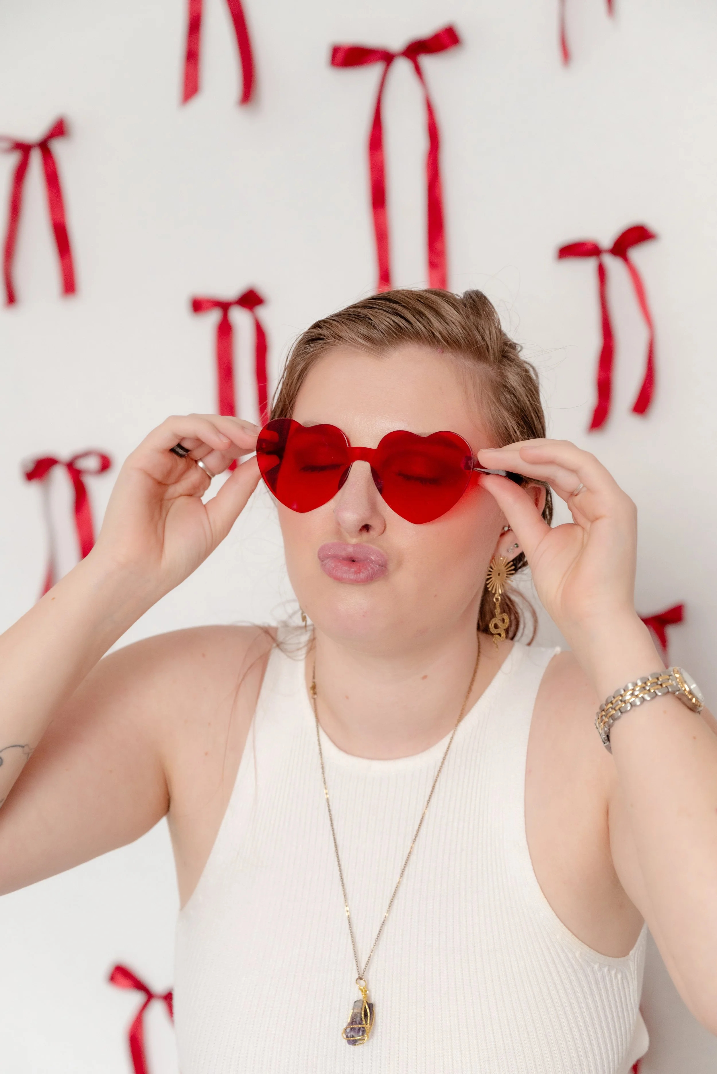 Close-up portrait of a woman wearing red heart-shaped sunglasses in The Alcove photography studio in Springville, Utah, featuring a white wall with red Valentine ribbon bows.