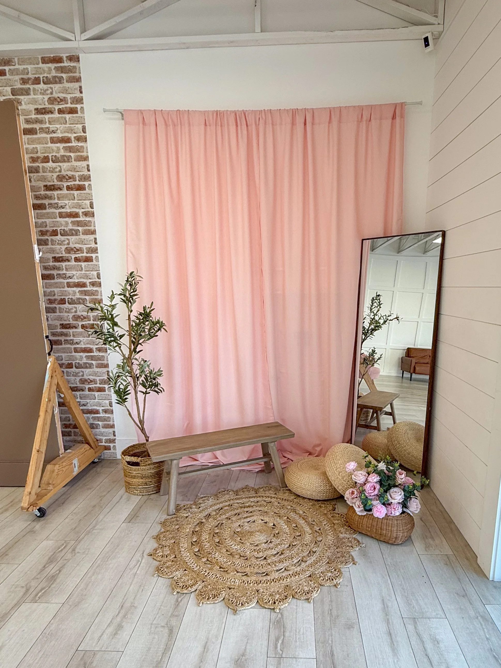 Romantic blush pink curtain backdrop inside a rentable natural light photography studio in American Fork, Utah. Styled with a wooden bench, floor mirror, wicker decor, florals, and greenery. Perfect for maternity photography, motherhood sessions, bra