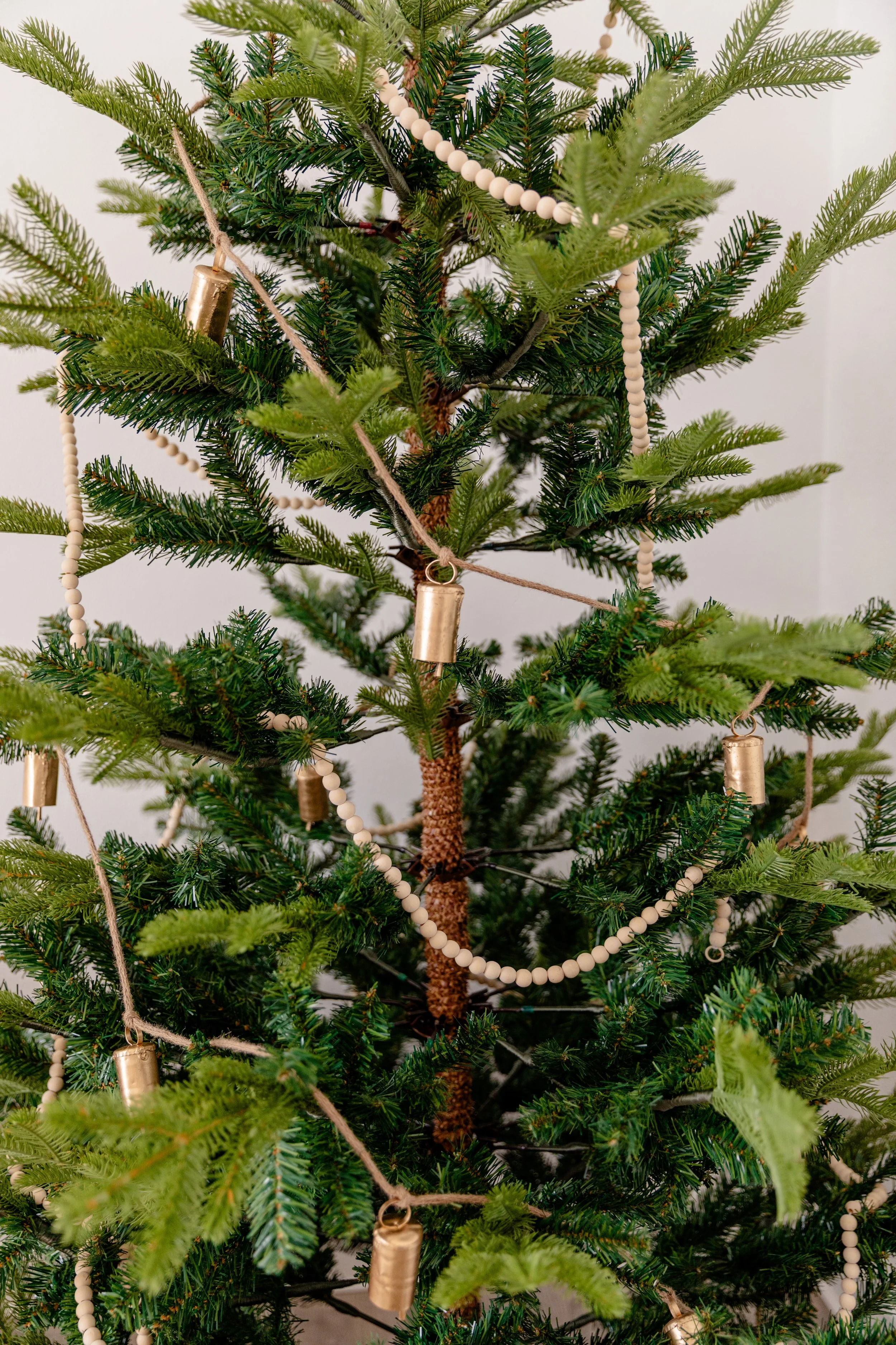 This close-up of the minimalist Christmas tree at The Alcove in Springville, Utah highlights its natural elegance — wooden bead garlands and small gold bells draped across lush green branches. The simple textures and neutral tones create a soft, orga