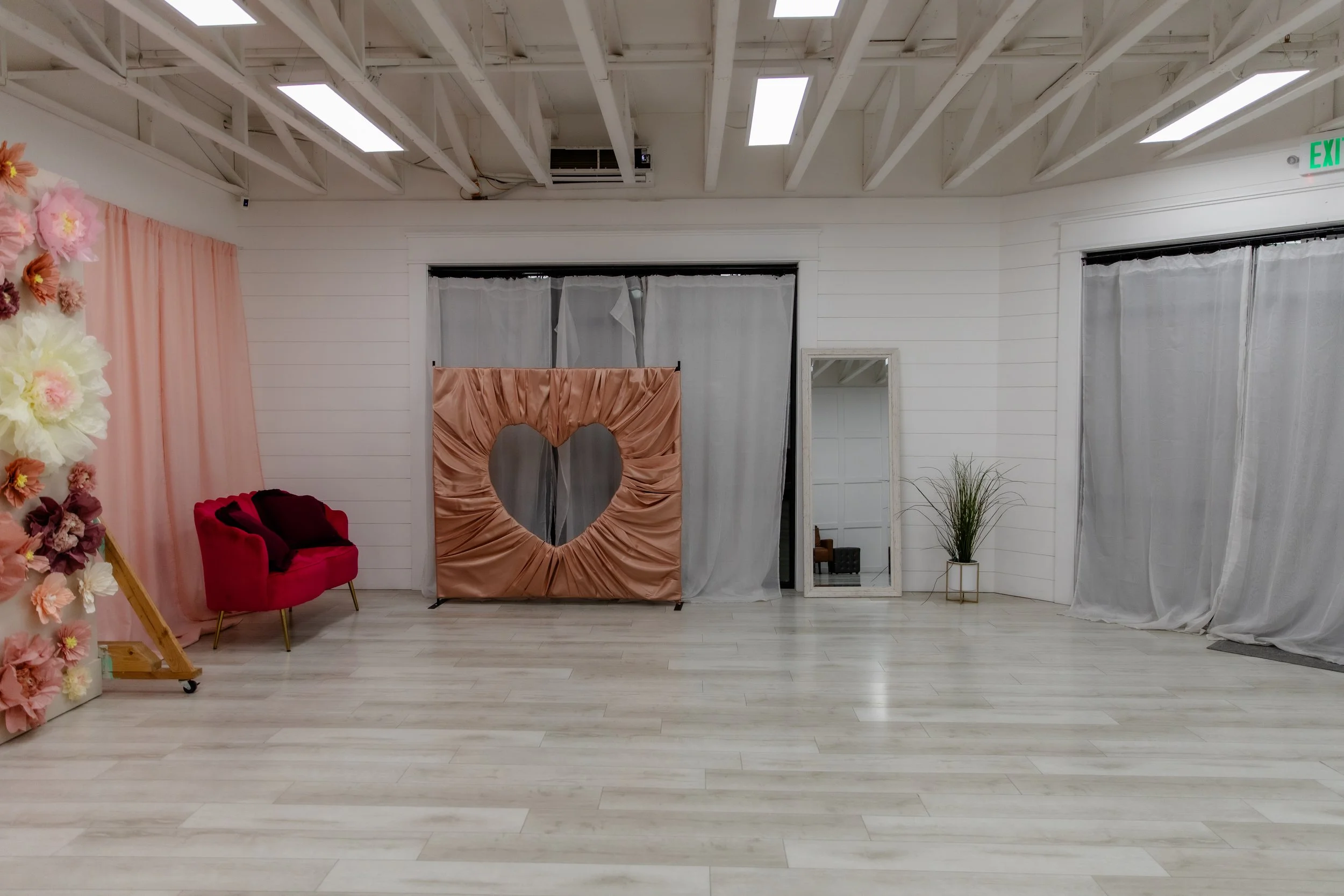 Side angle view of Studio A at Varsa Studios American Fork showing Valentine’s Day photo setups, heart backdrop, floral wall, pink curtain, and natural light windows for portrait photography and content creation.