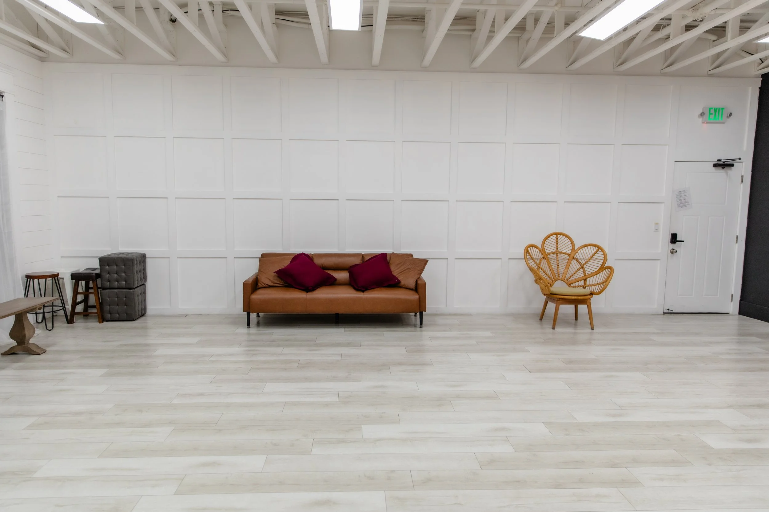 Wide view of Studio A at Varsa Studios American Fork featuring a white paneled wall, brown leather couch, wicker chair, and open floor space, perfect for photographers and content creators needing a versatile natural light studio.