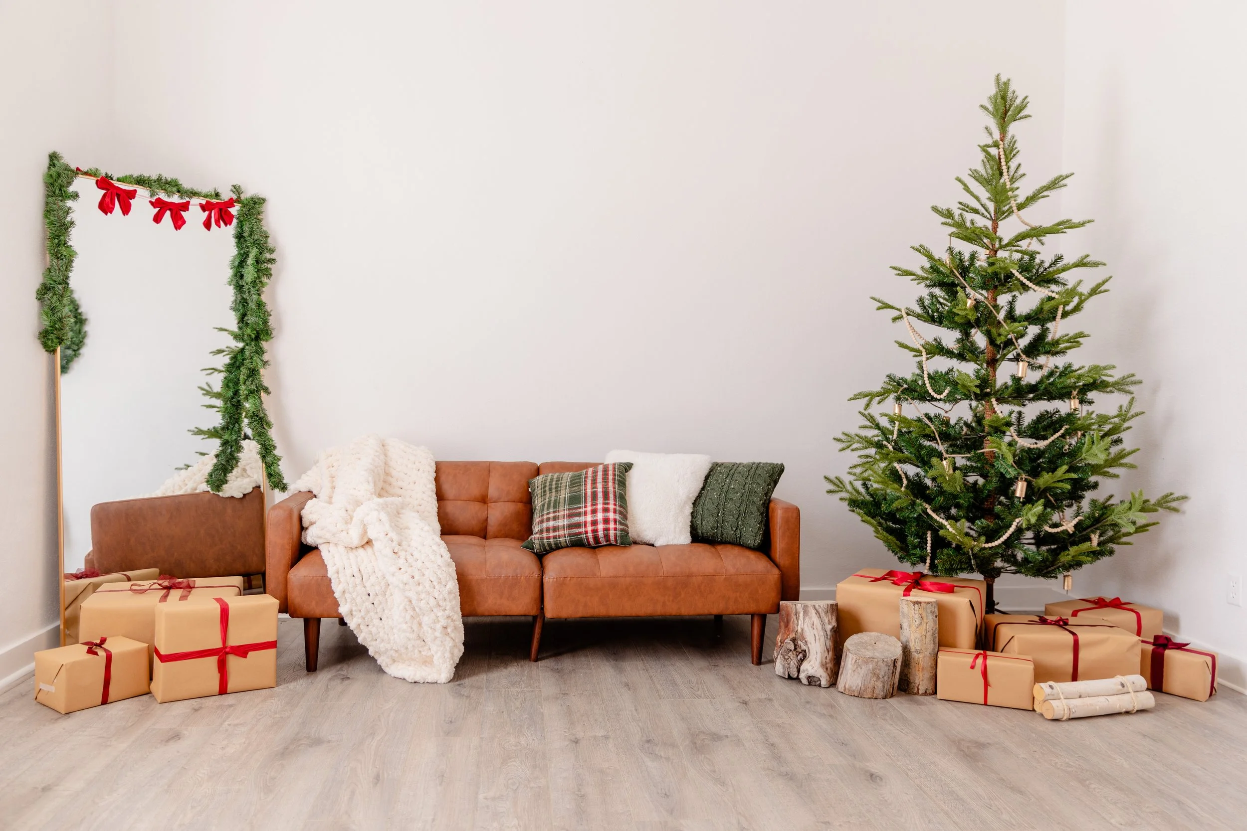 Step into a cozy, modern Christmas scene at The Alcove in Springville, Utah, part of Varsa Studios. This setup features a warm brown leather sofa accented with chunky knit blankets, plaid and textured pillows, a minimalist tree with gold accents, and