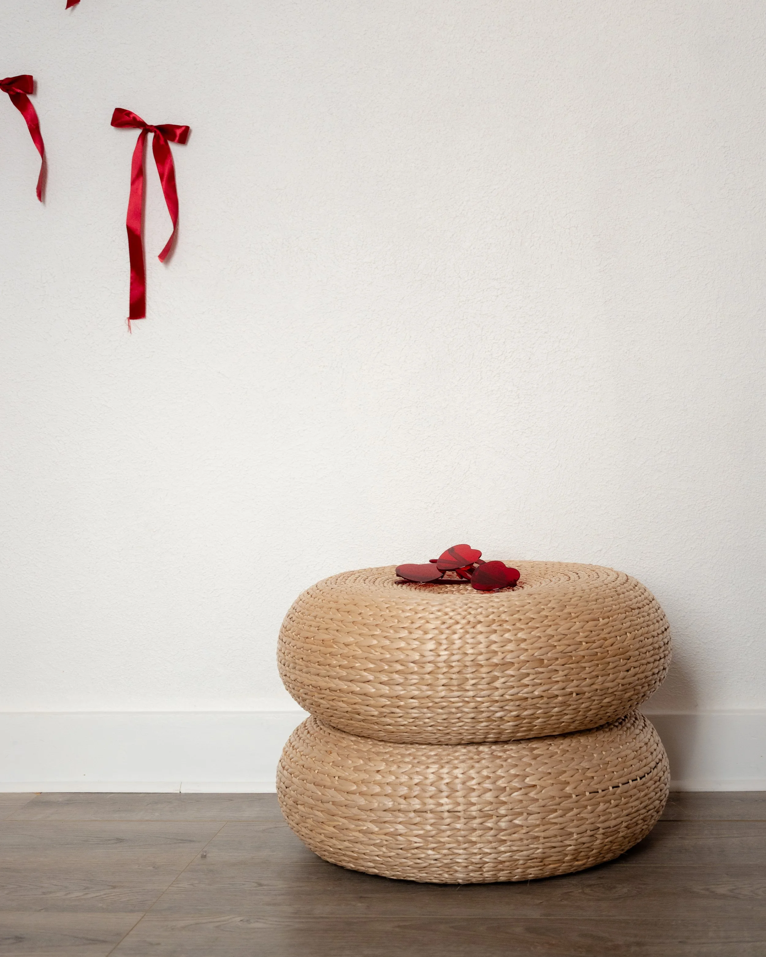 Woven rattan ottomans styled beneath a white wall with red ribbon accents in The Alcove photography studio in Springville, Utah.