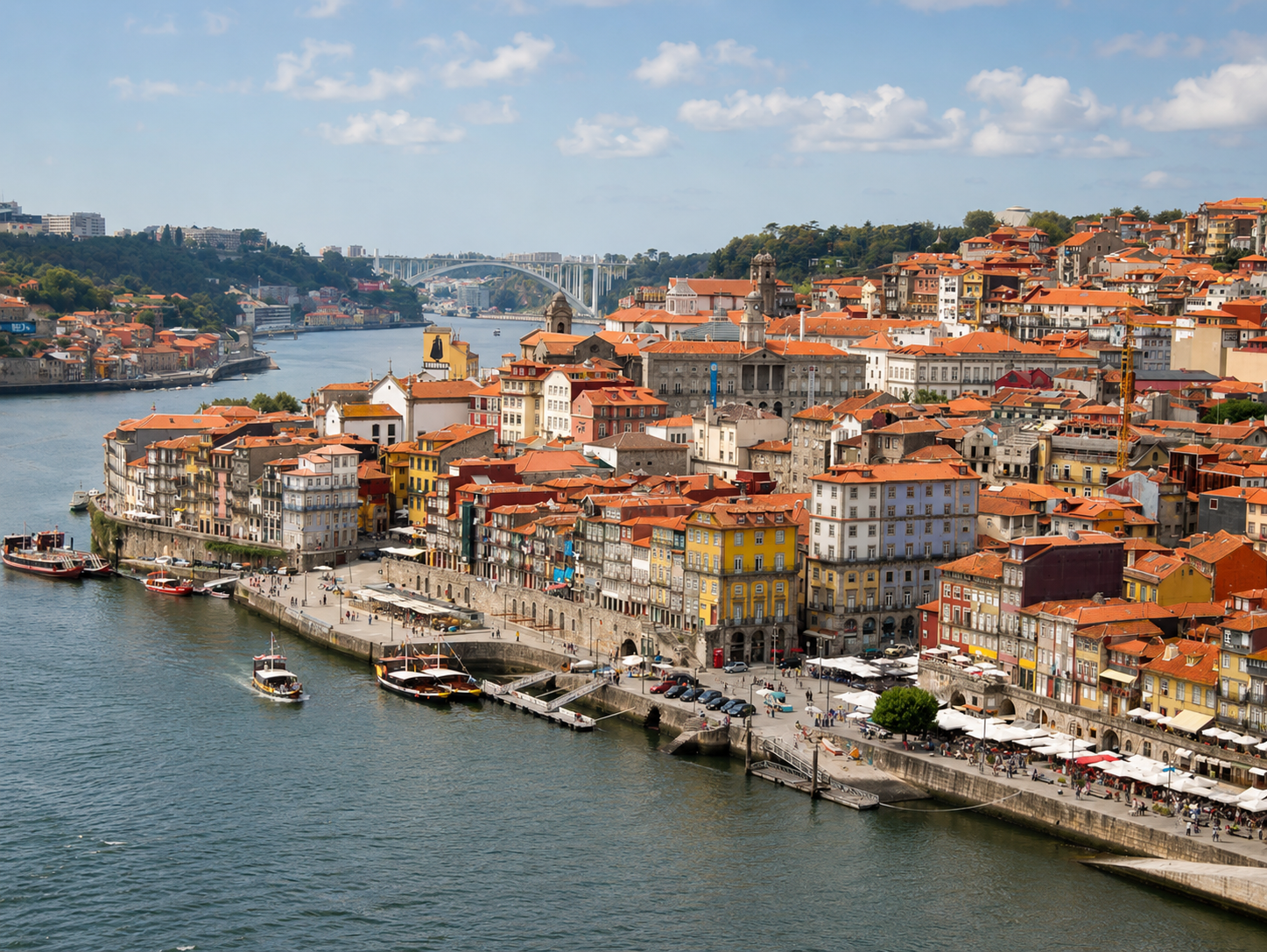 Ribeira waterfront in Porto, Portugal, with colorful historic buildings, boats on the Douro River, and the Arrábida Bridge in the distance.