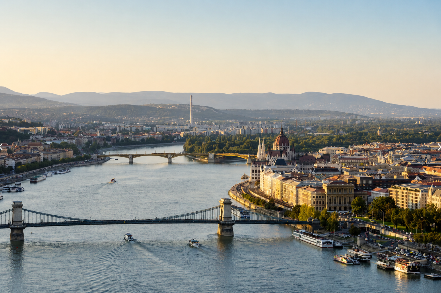 Panoramic view of Budapest, Hungary, featuring the Danube River, Széchenyi Chain Bridge, Hungarian Parliament Building, and historic Pest skyline at golden hour.